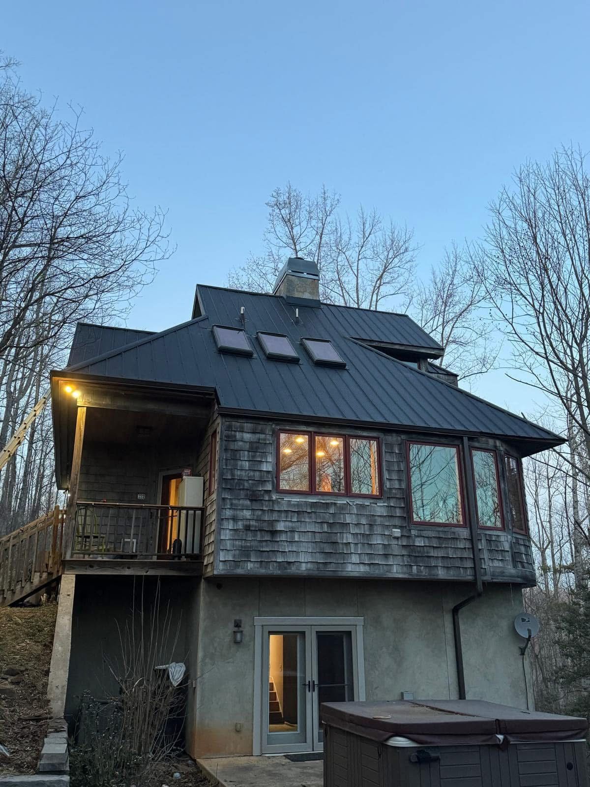 Two-story wooden house with a dark metal roof. A deck, windows, and a hot tub are visible.
