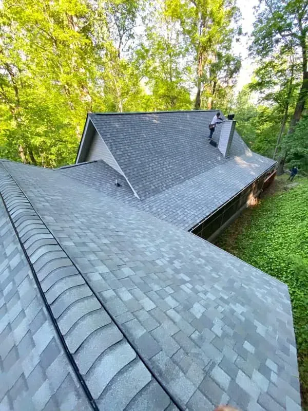 Gray shingle roof, person working on the peak, surrounded by trees.