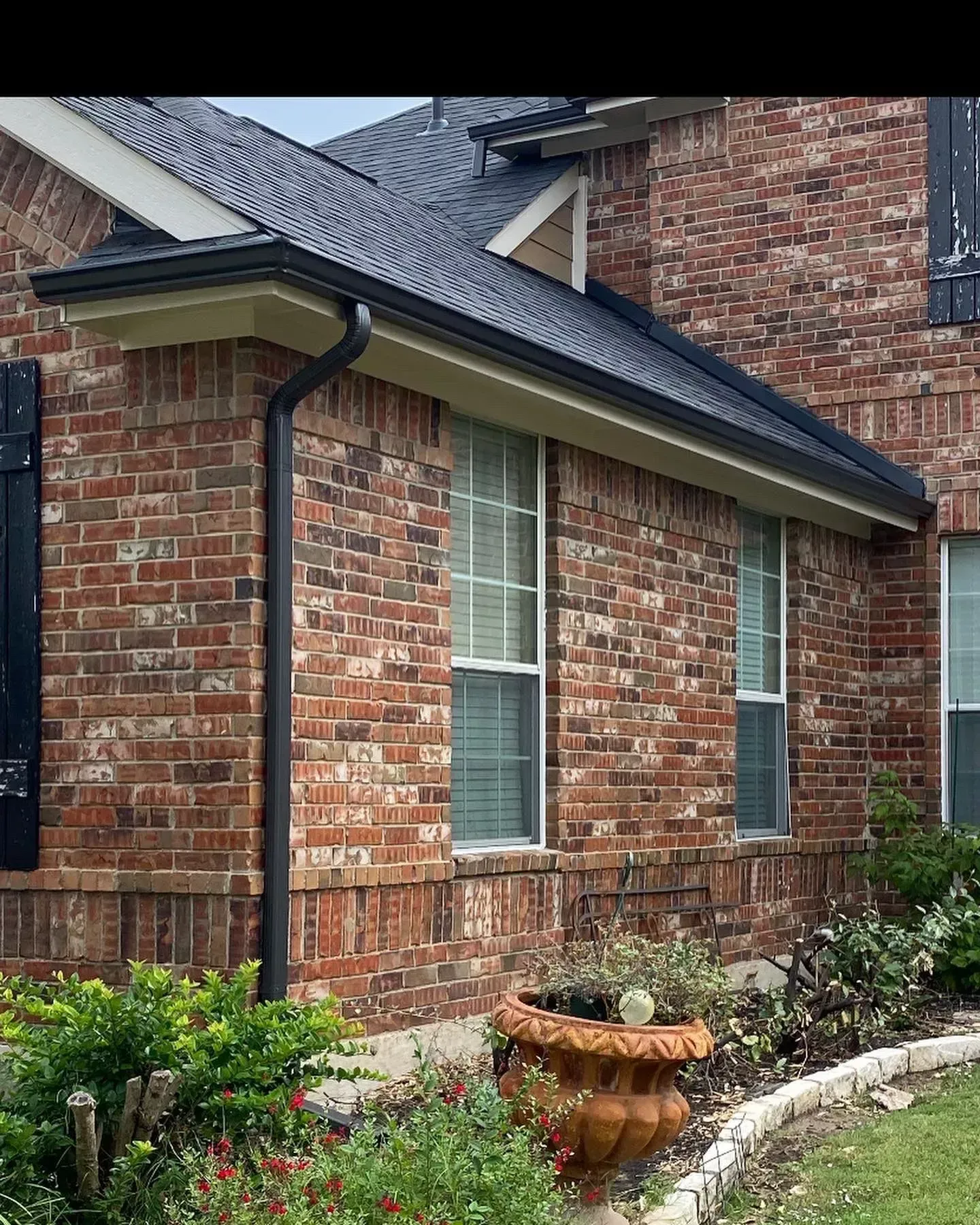 Red brick house with black gutters, windows, and roof. Landscaping in front.