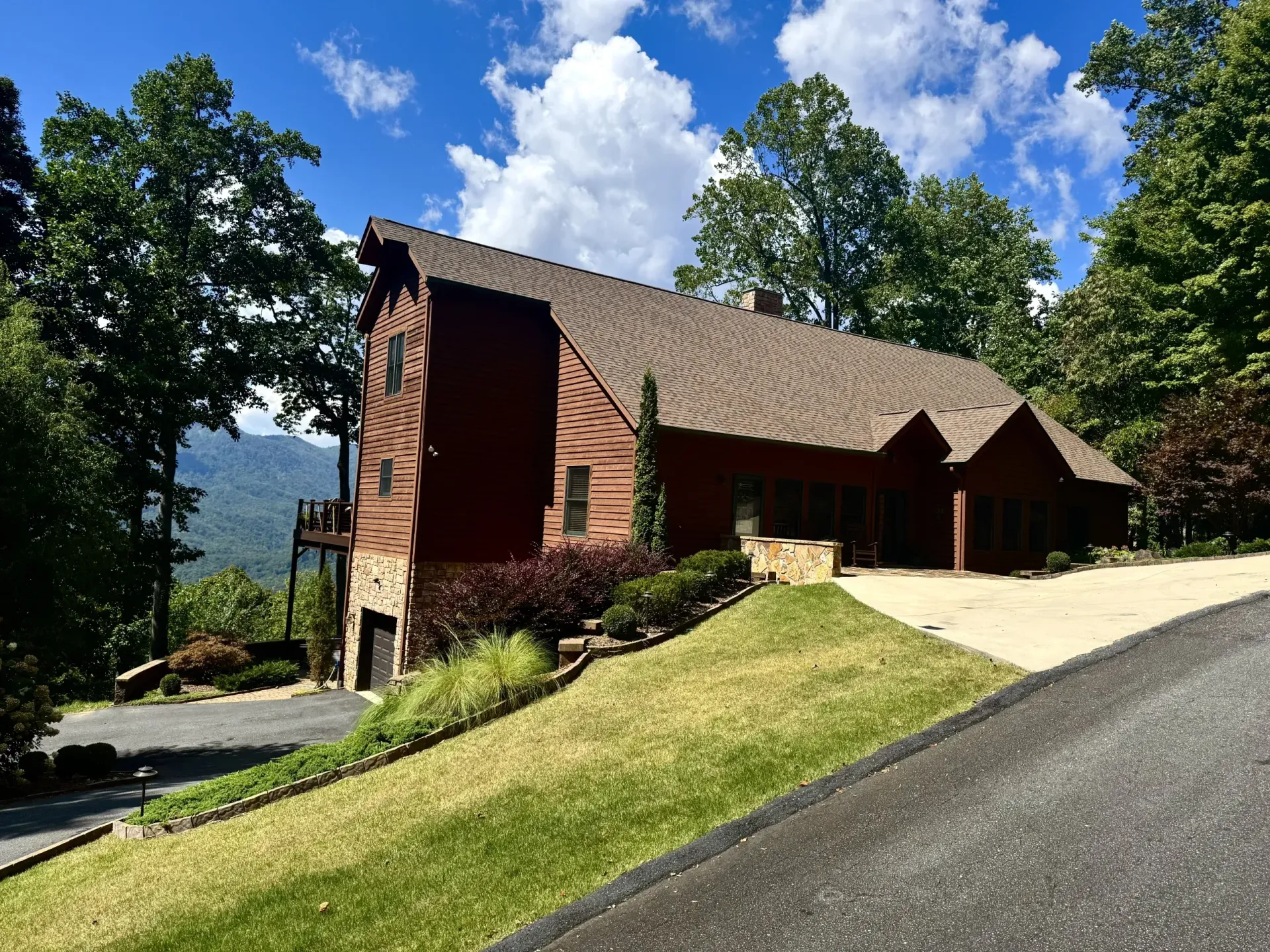 Brown wooden house on a hillside with a driveway, surrounded by trees and a cloudy sky.