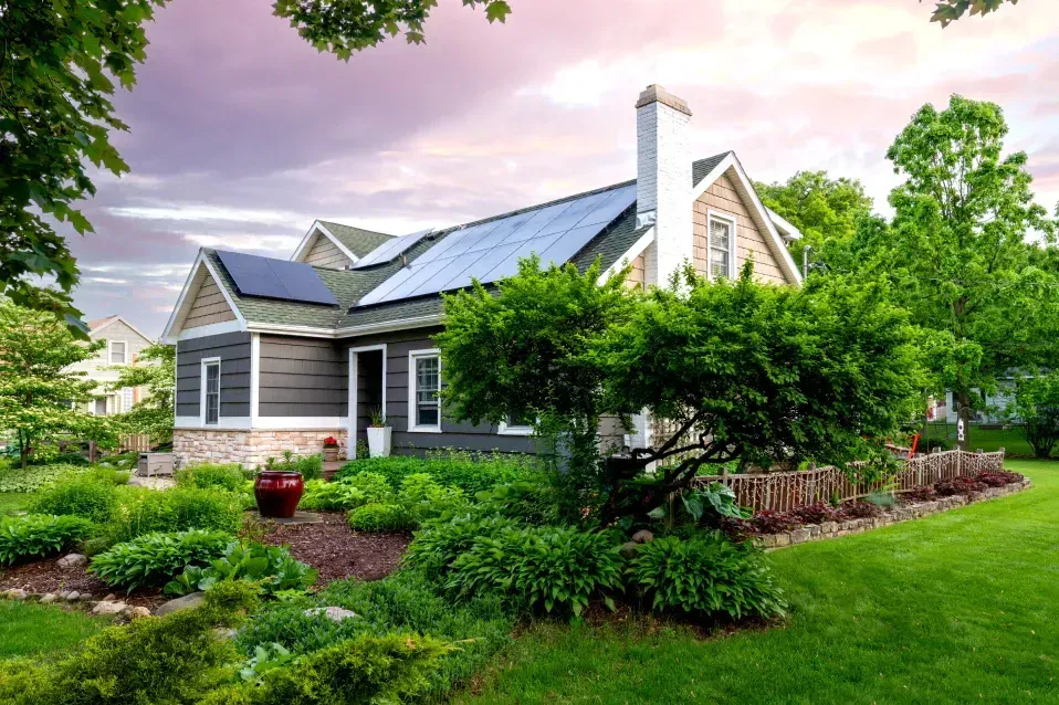 House with solar panels on roof, surrounded by green garden and trees.