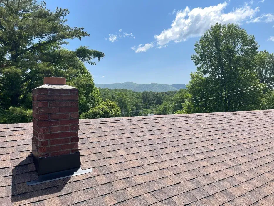 Brick chimney on a brown shingled roof with a scenic view of green trees and mountains under a blue sky.