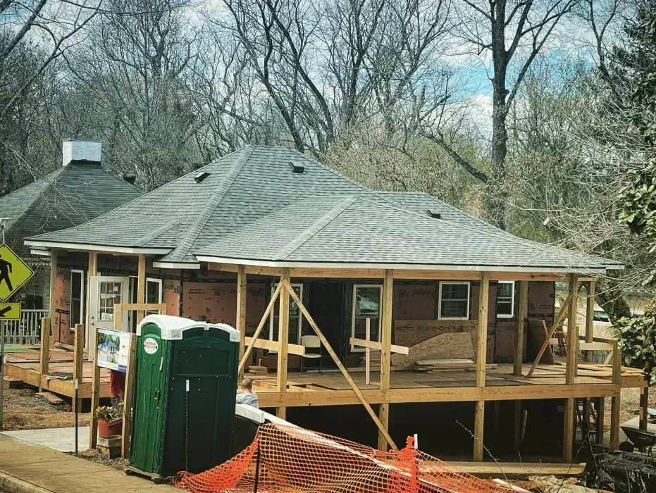 A house under construction with a wraparound porch; a green portable toilet sits in front.