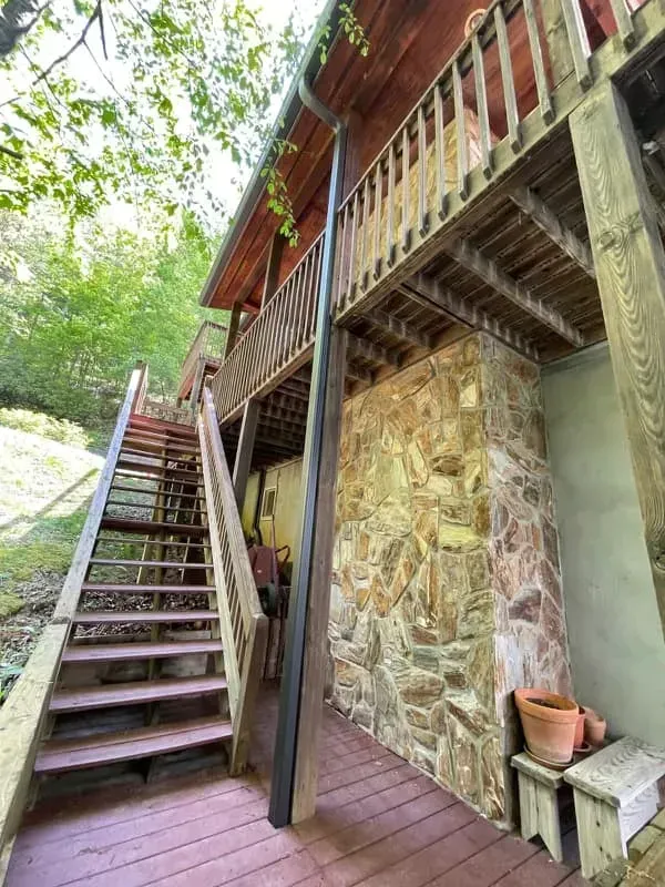 Wooden staircase leading to a two-story house with a stone foundation, surrounded by trees.