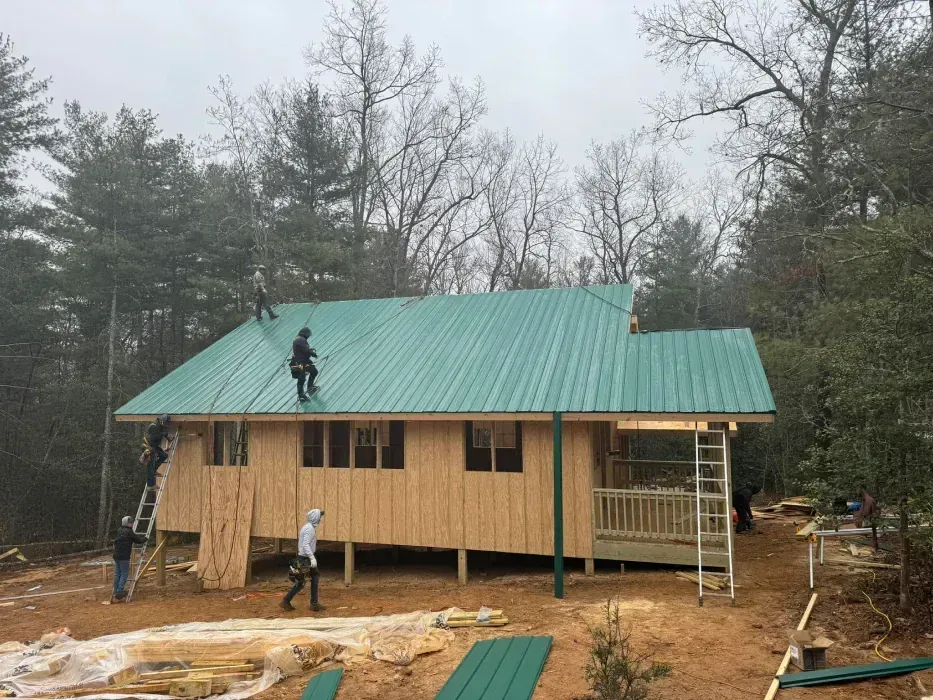 Construction of a house with a green metal roof. Workers on roof and at walls, set in a wooded area.