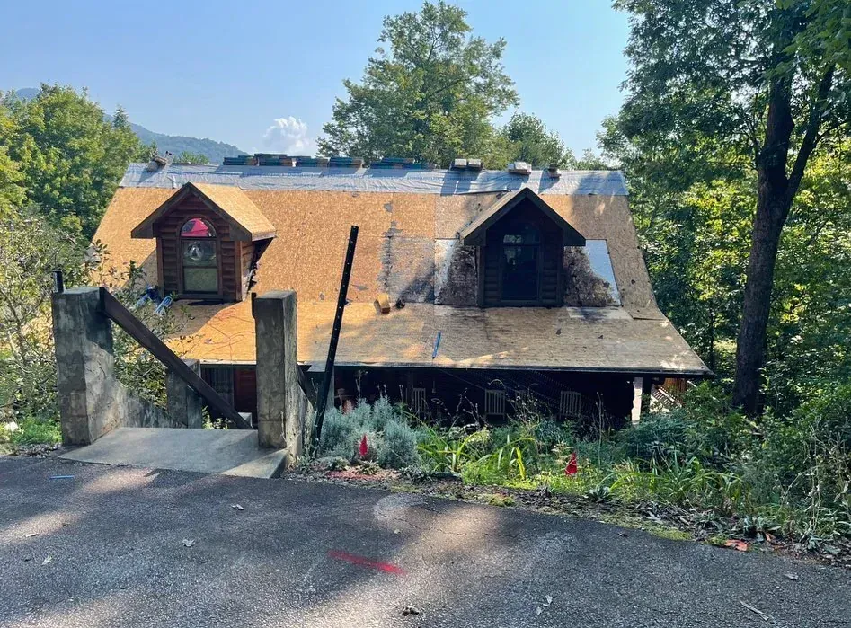 Cabin with partially replaced roof, two dormers, surrounded by trees, viewed from a road.