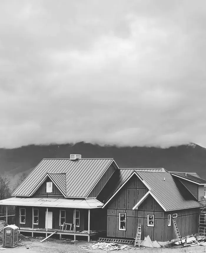 Black and white photo of a house under construction with a metal roof, cloudy sky, and mountains in the background.
