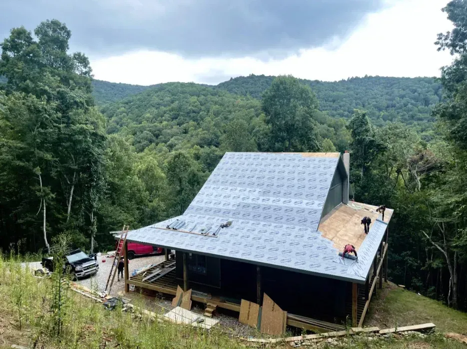 Cabin under construction on a hillside, surrounded by trees. Workers on roof. Cloudy sky.