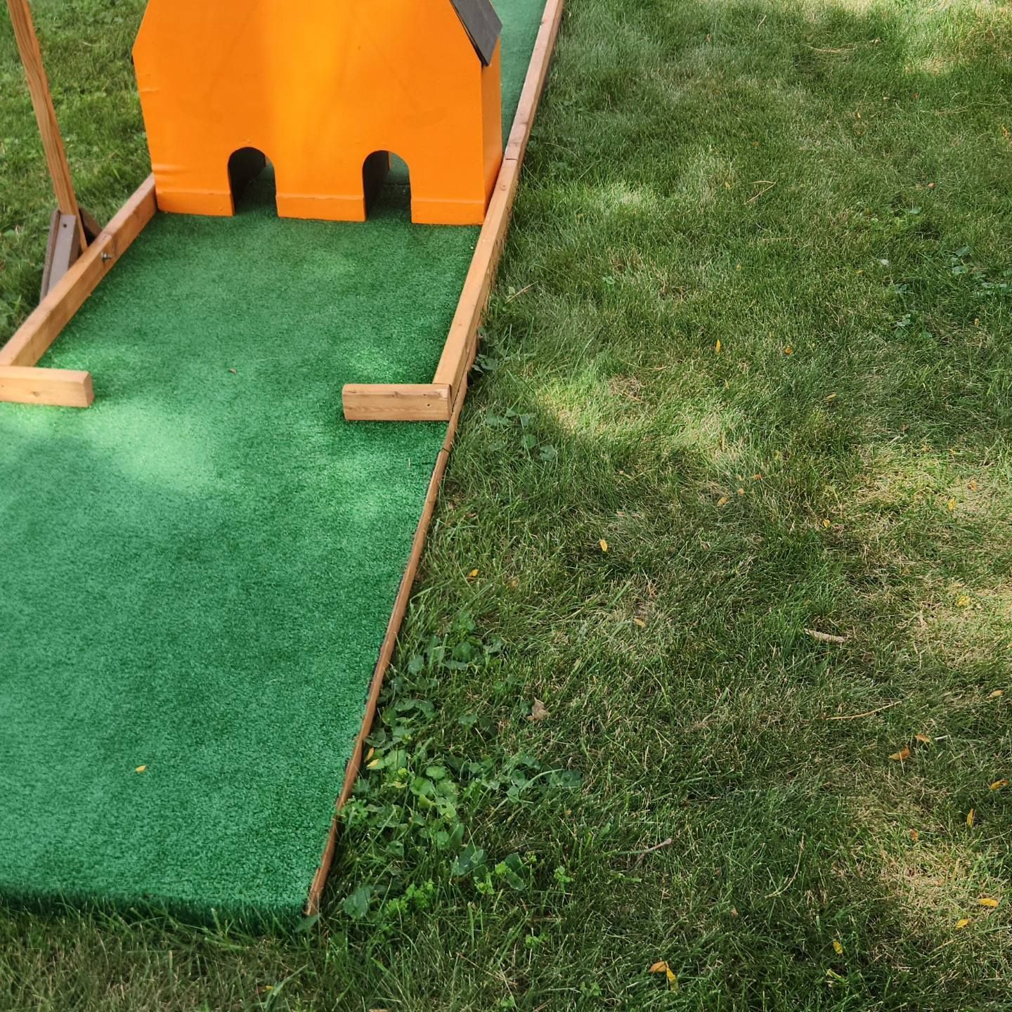 Miniature golf course, green turf, orange obstacle, next to grass.