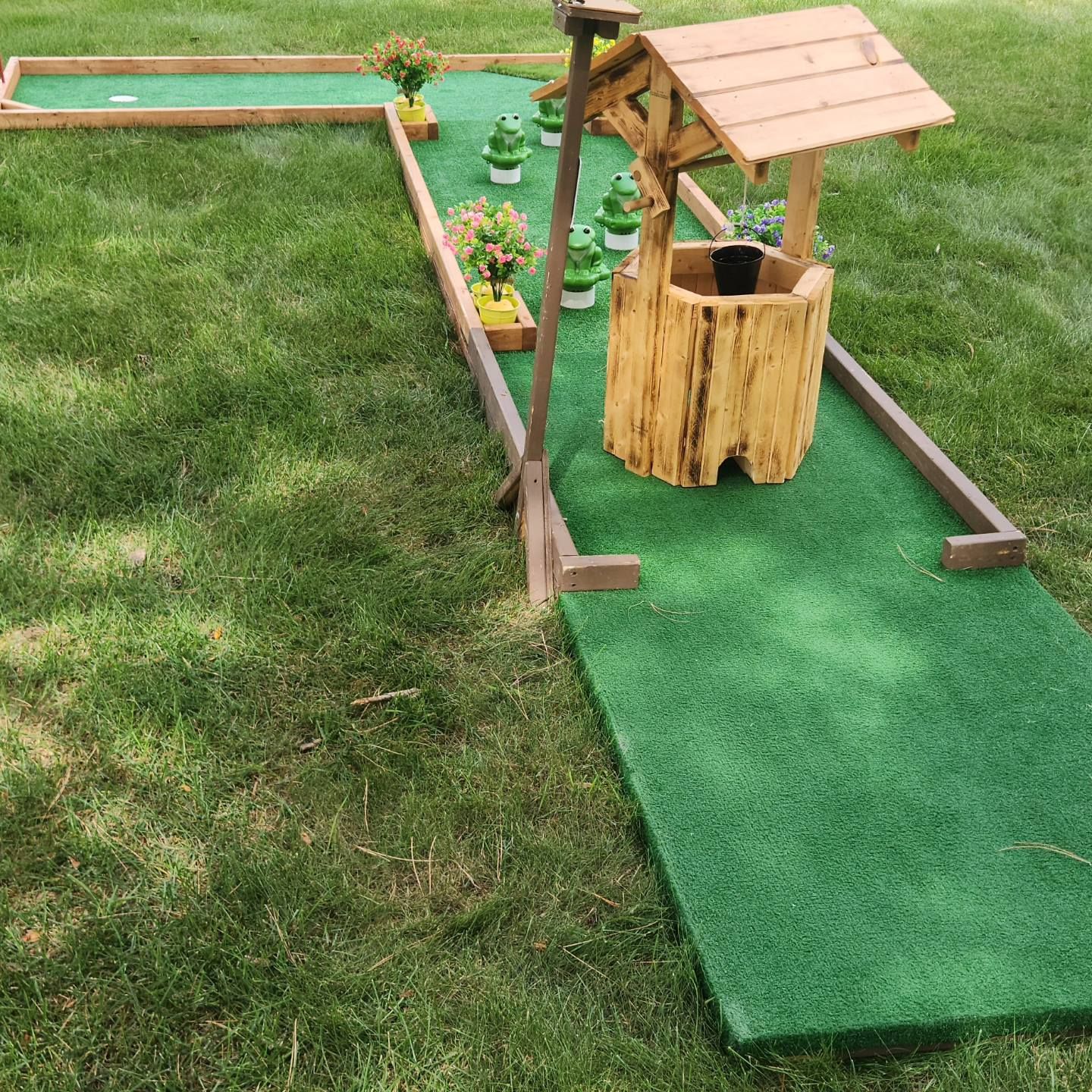 Mini golf course: green turf, wooden well, frog statues, flowers, and a grassy lawn.