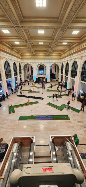 Miniature golf course set up inside a grand, ornate building with high ceilings. People playing.