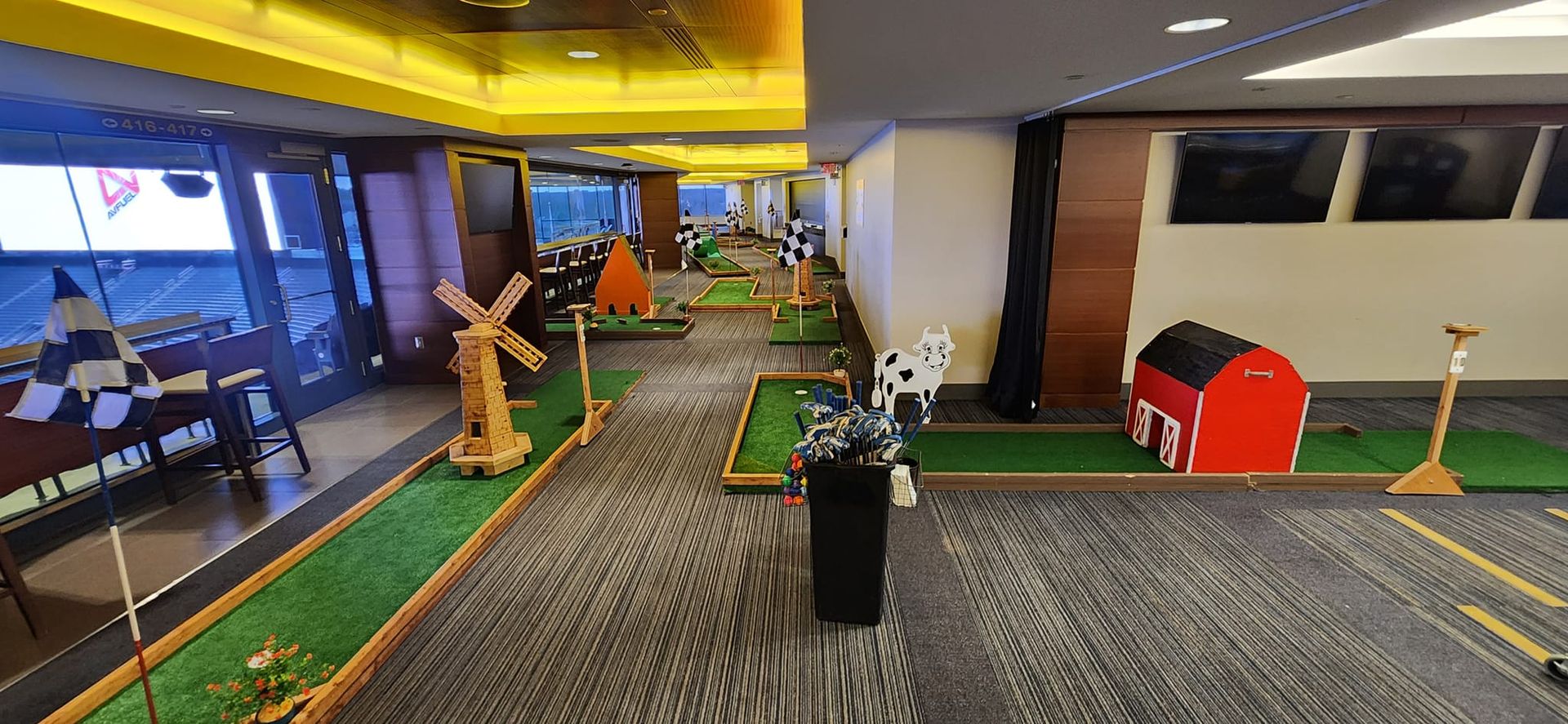 Indoor mini golf course, featuring windmill, cow, and red barn obstacles on green turf.