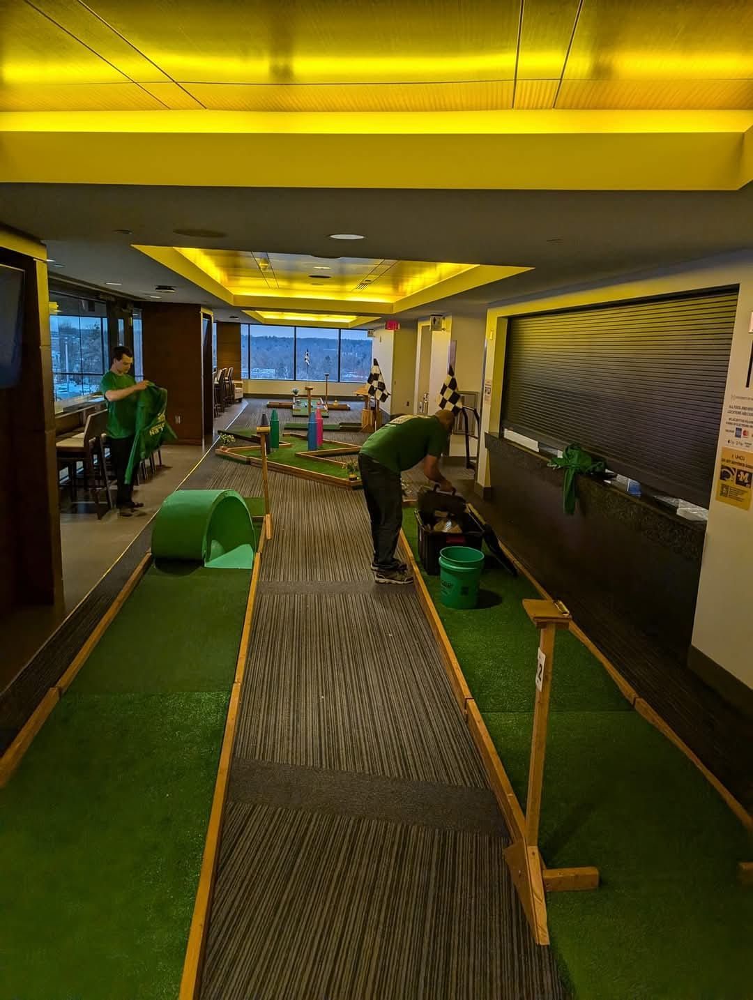People playing miniature golf indoors with green felt courses and a cityscape view.