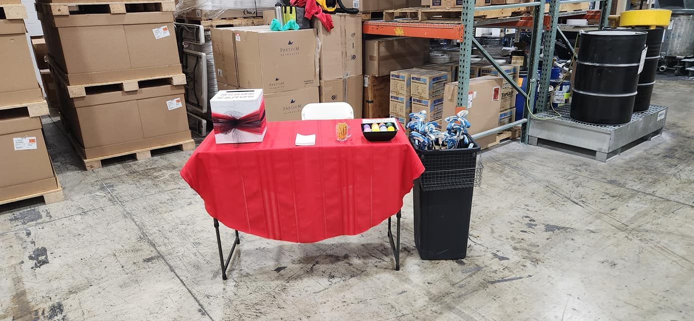 A red-covered table with items sits in a warehouse. A trash can is beside it. Cardboard boxes in the background.