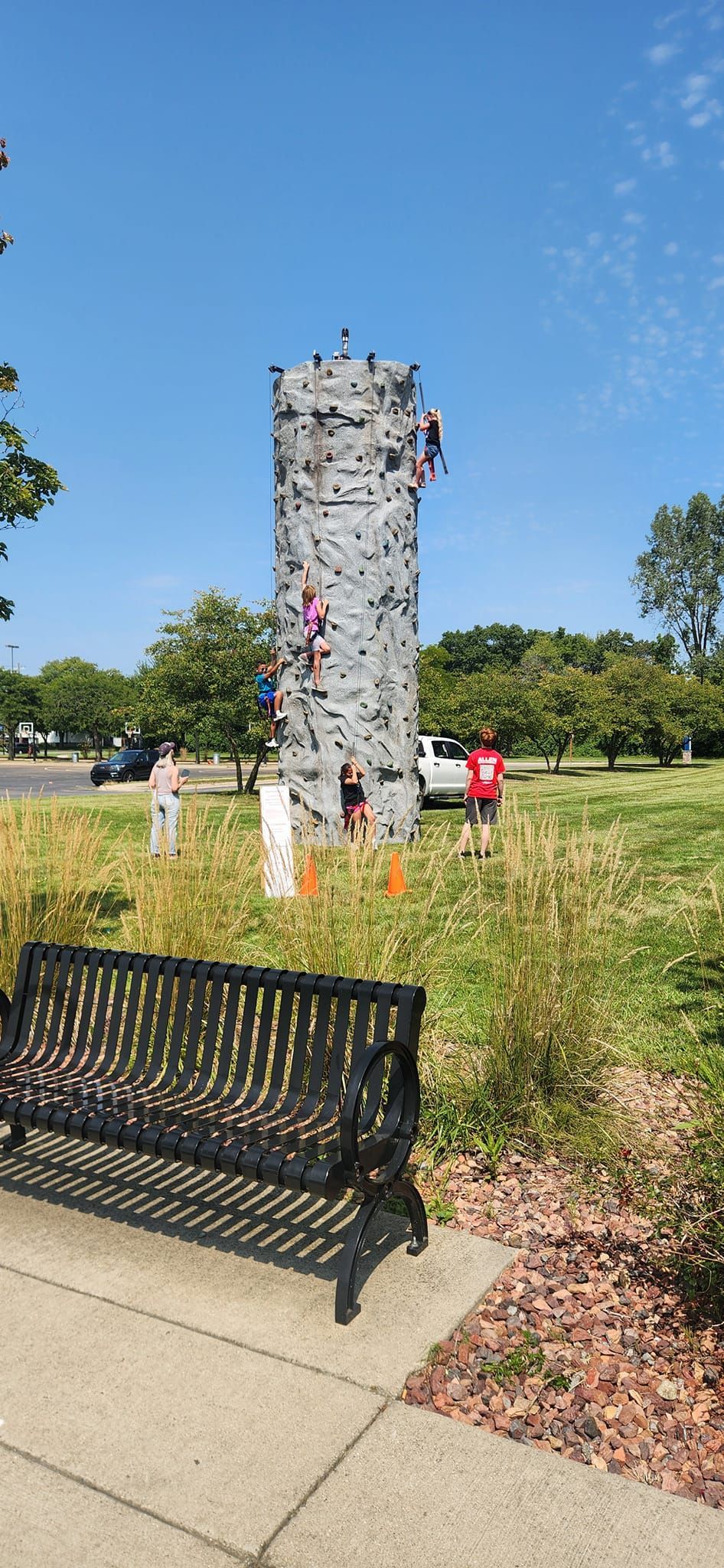 People climbing a gray rock climbing tower in a park on a sunny day. A bench is in the foreground.