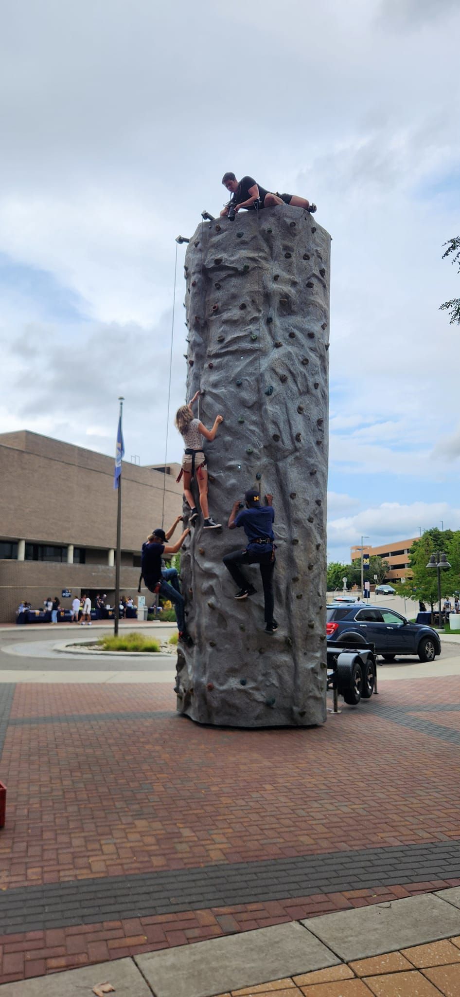 People climbing a tall outdoor rock climbing wall. Set in a brick paved square.