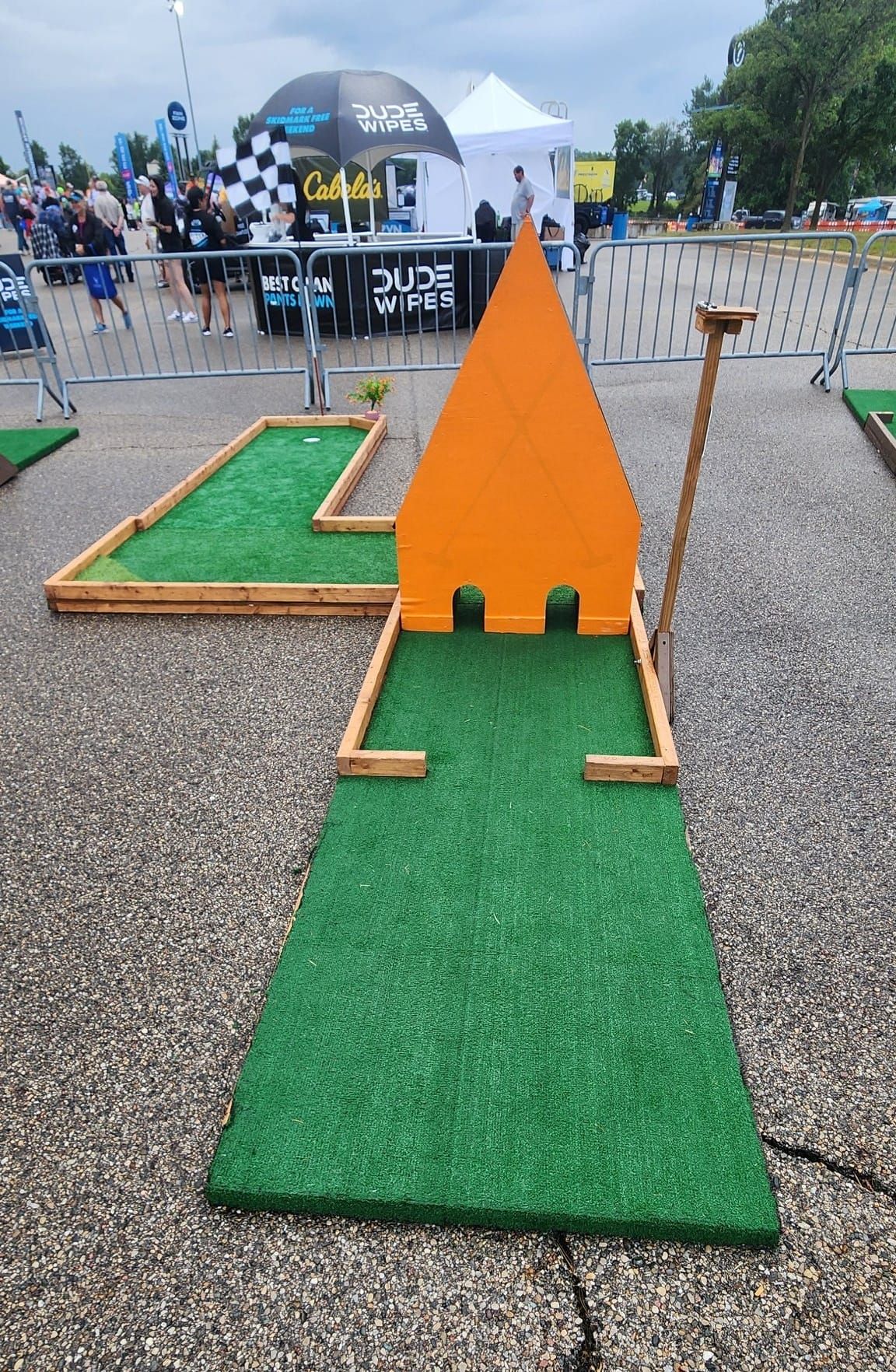 Mini golf hole with orange obstacle; green turf, wooden border, gray pavement, event setting.
