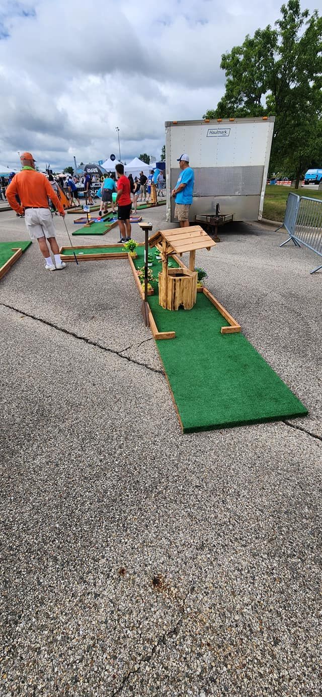 People playing mini golf on a concrete surface. One course has a wooden well. Cloudy sky.