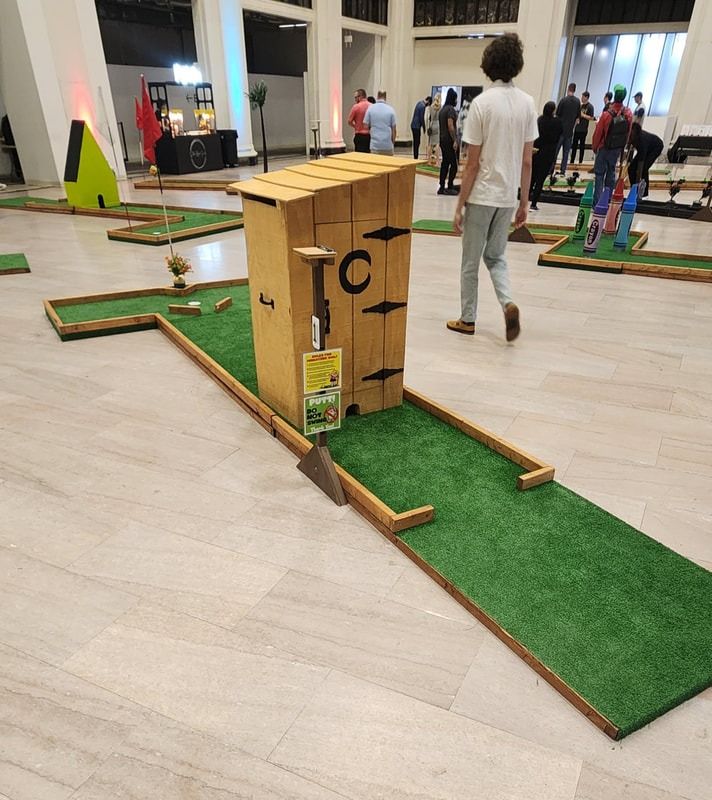 Indoor mini golf course with a wooden outhouse obstacle. People play in a brightly lit space.