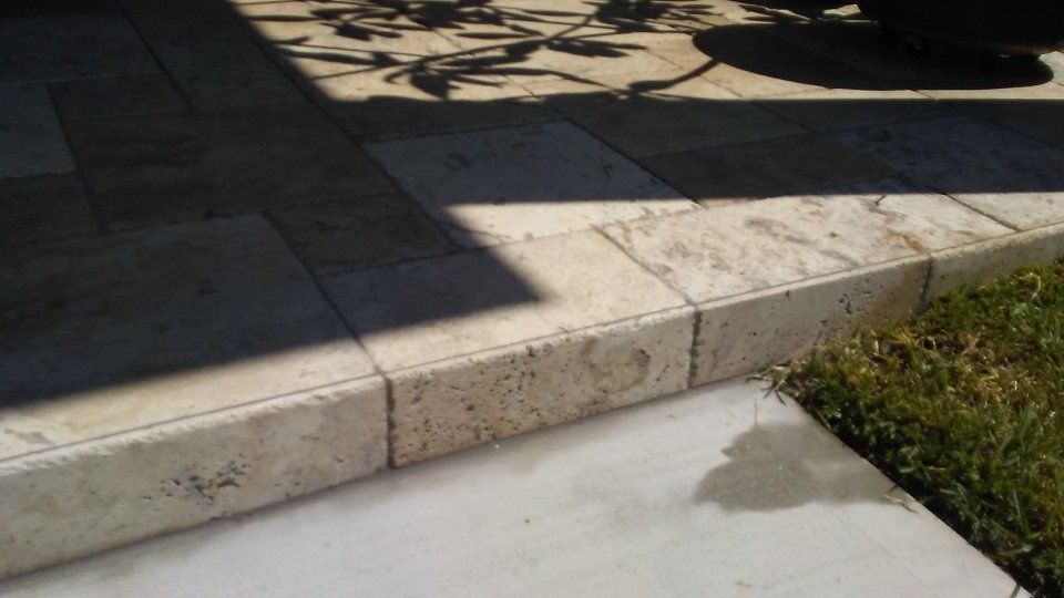 Travertine stone patio edge next to grass and concrete. Sunlight and shadows.