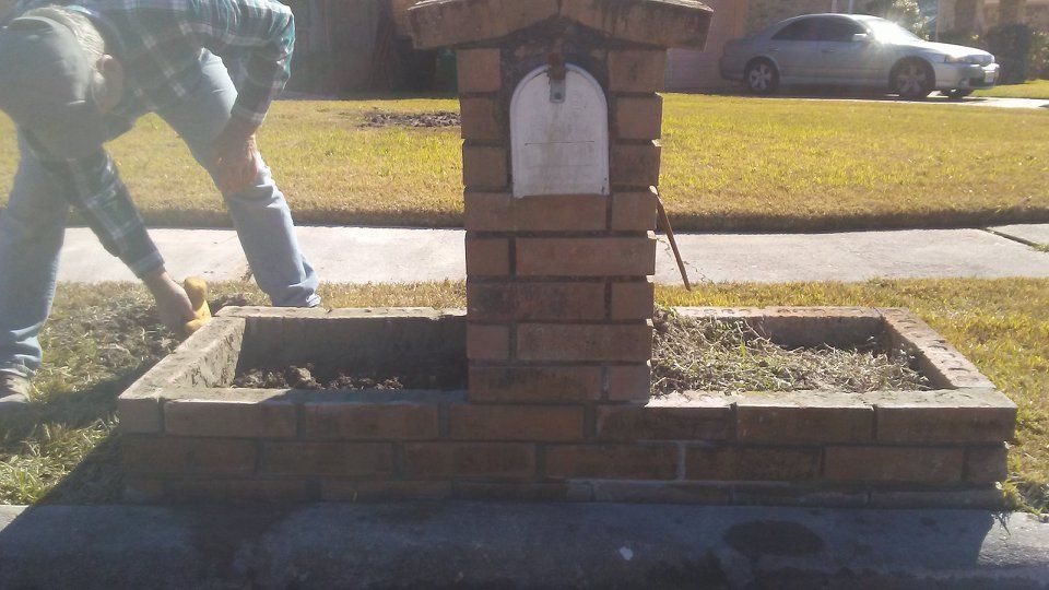 Man tending a brick mailbox with a flower bed.  A car sits in the background on a sunny day.