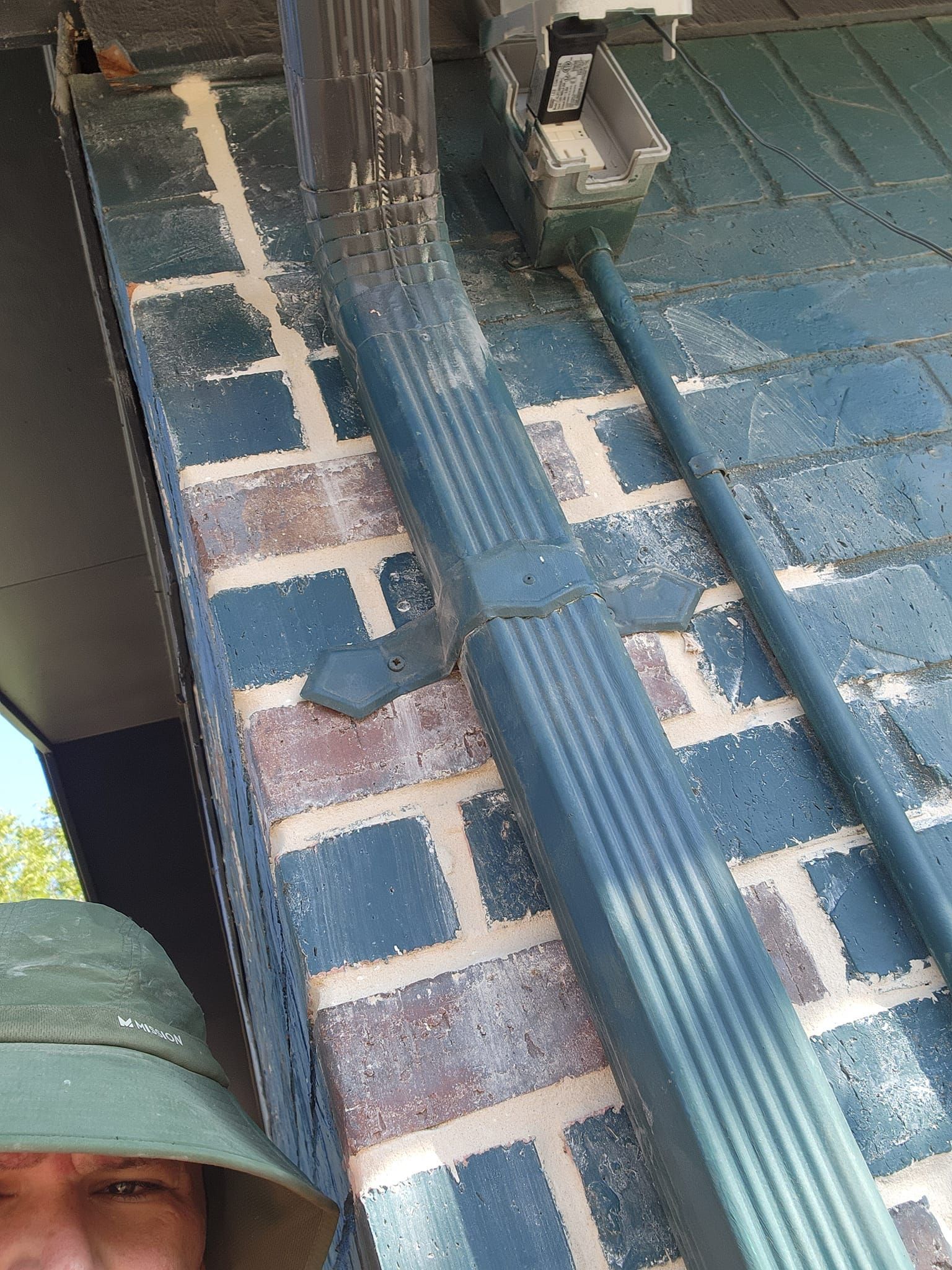 Blue brick wall with blue downspout and gutter, part of a face visible, wearing a green hat.