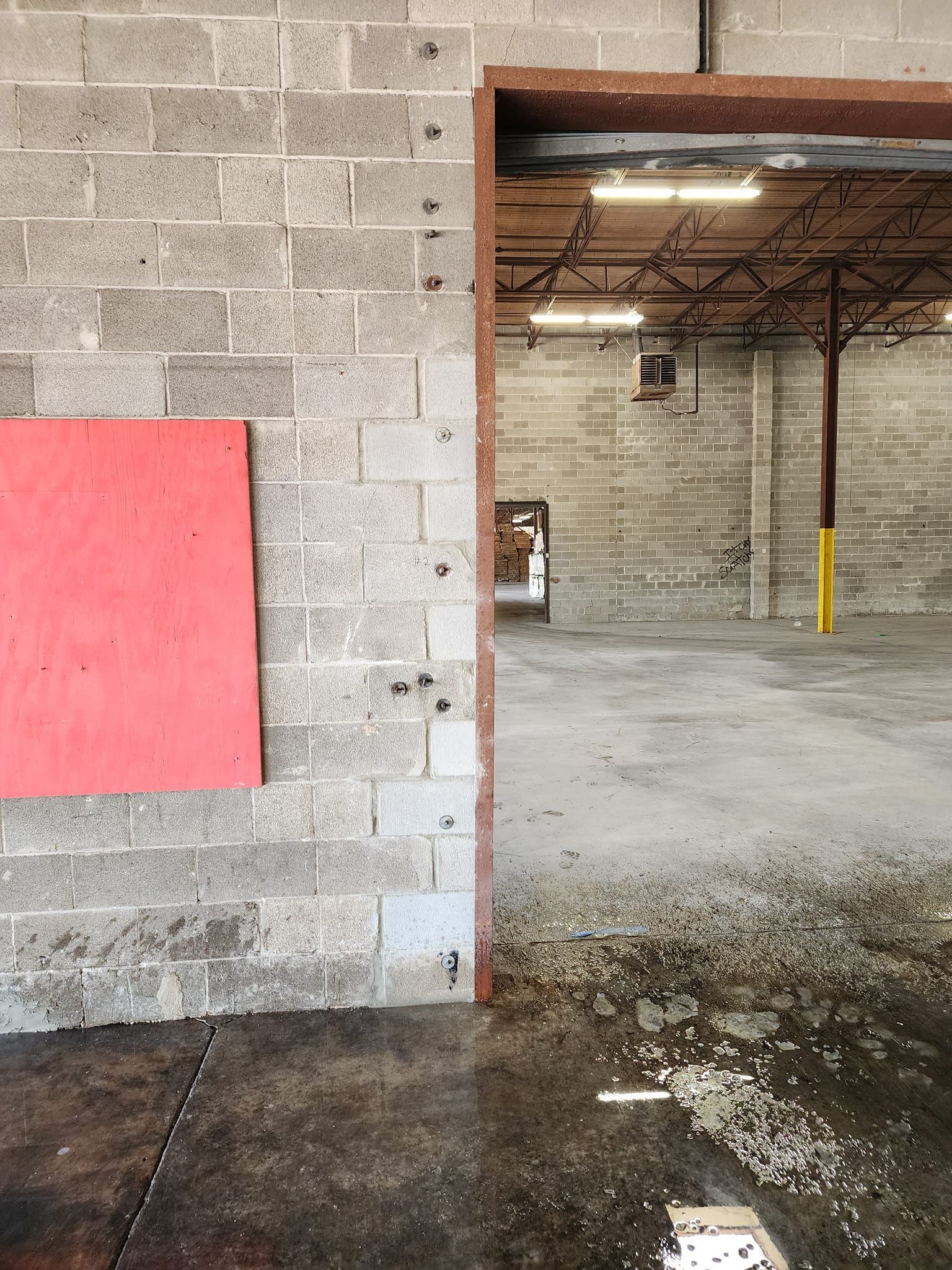 Interior of a warehouse with a concrete floor, concrete block walls, and open doorway. A red panel is attached to a wall.