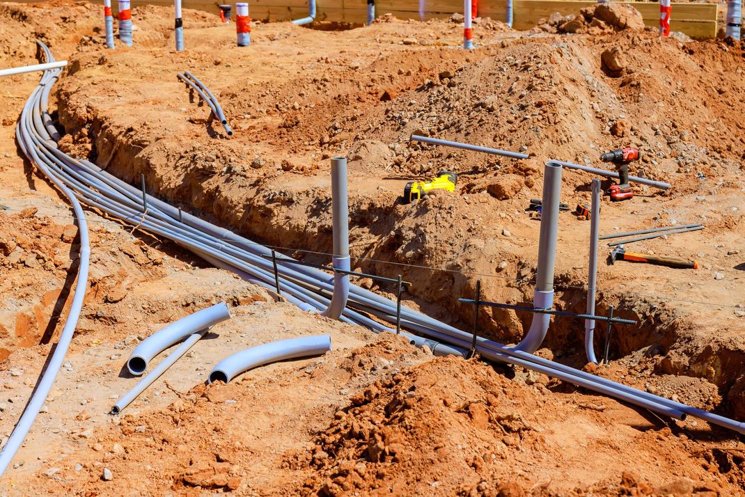Un cantiere edile caratterizzato da tubi e condotti elettrici grigi posati in una trincea in un terreno bruno-rossastro.