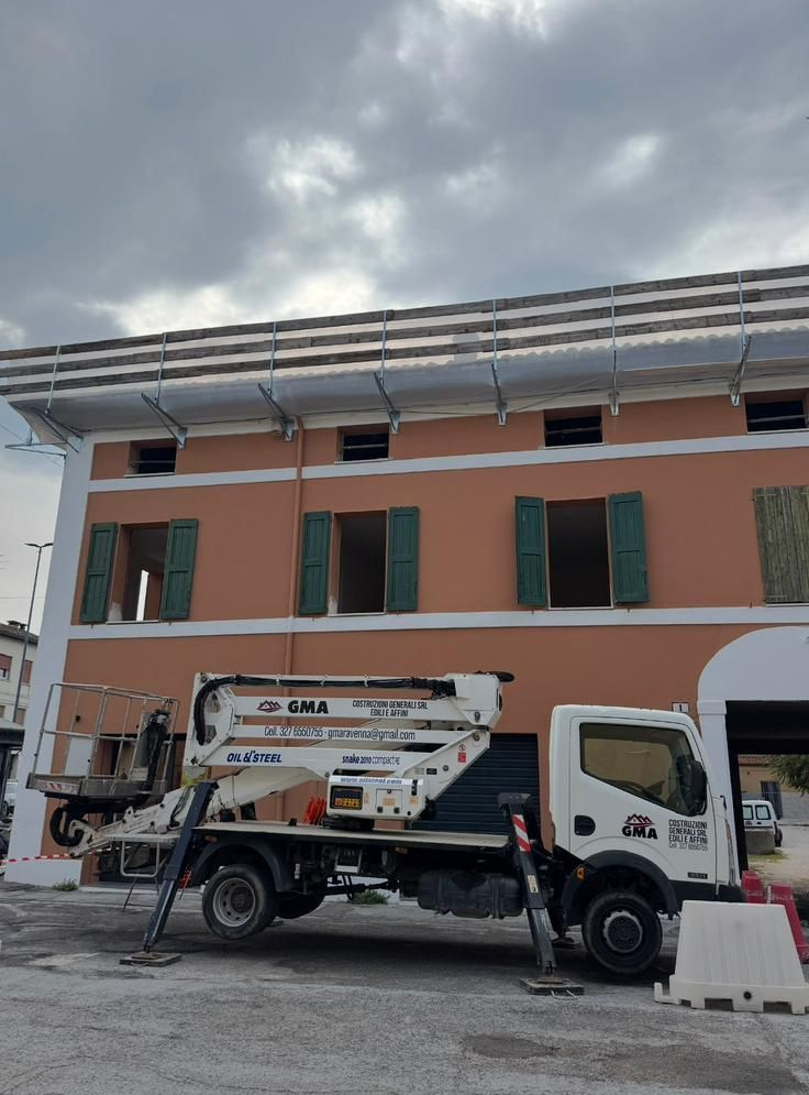 Una piattaforma aerea di lavoro bianca montata su un camion è parcheggiata di fronte a un edificio di tre piani color pesca.