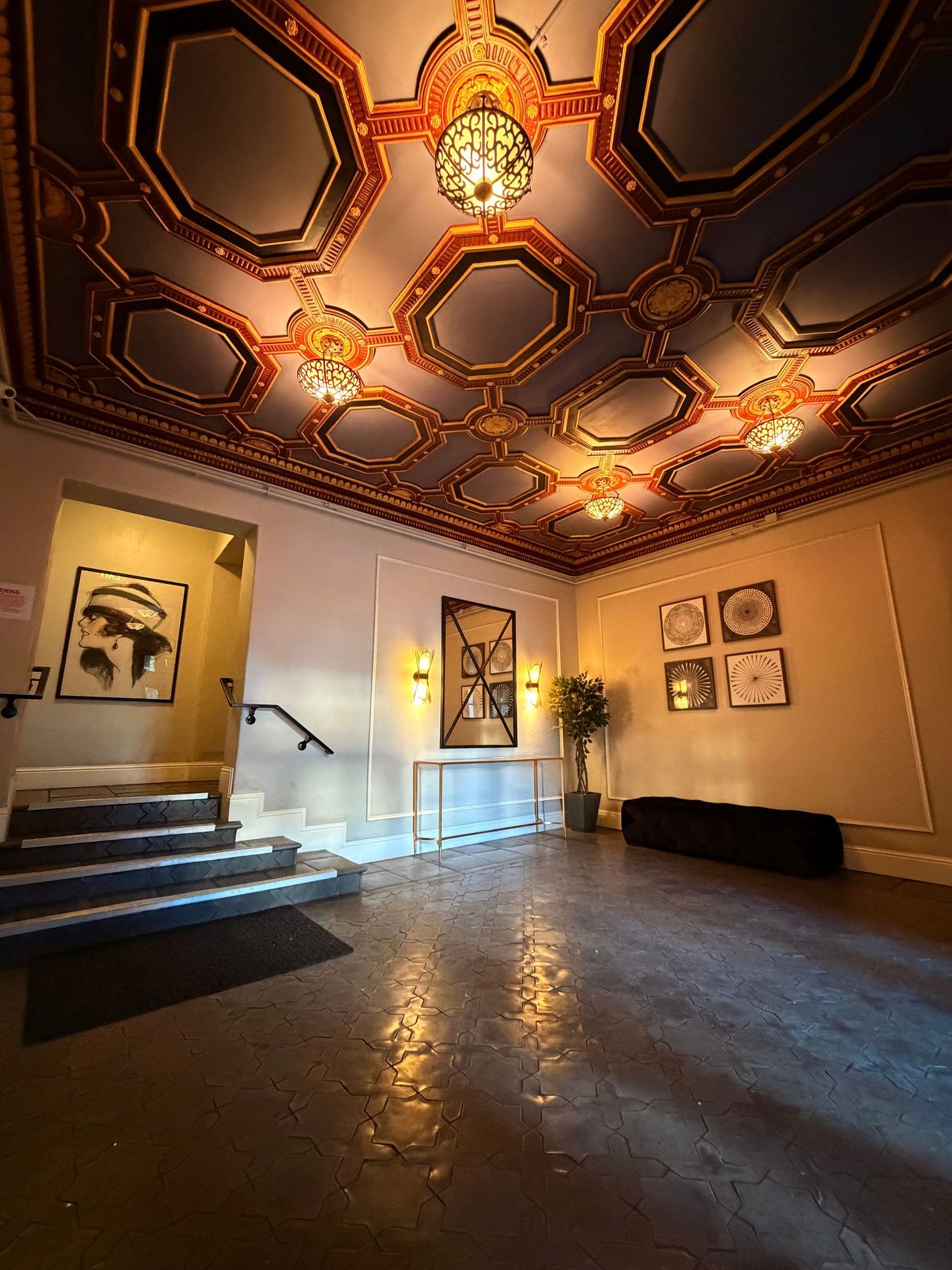 Ornate ceiling of an entryway; gold, blue, and brown. Artwork and stairs are visible.