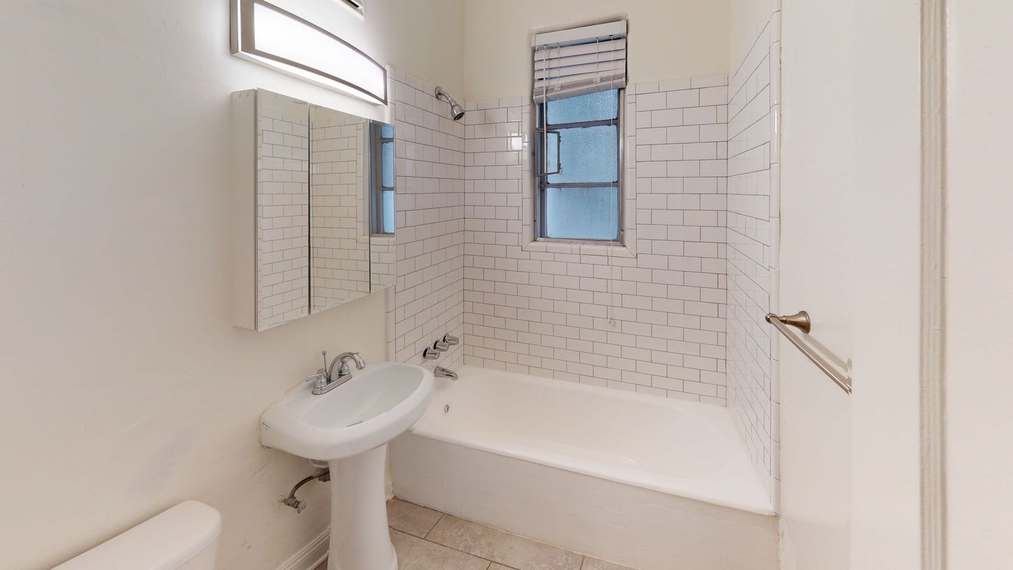 Small, white bathroom with a sink, toilet, tub, and window with a blind. Tile on the walls.