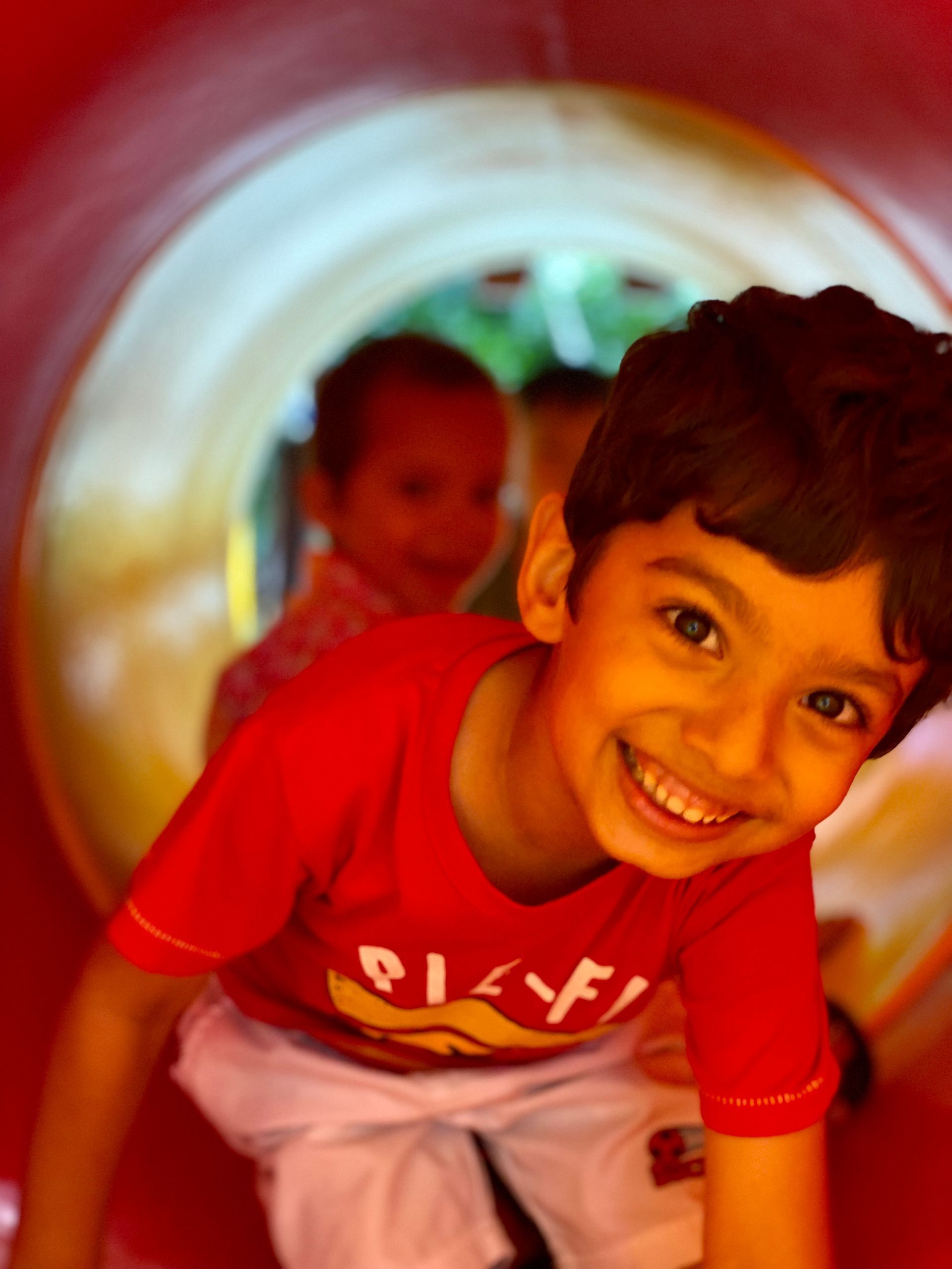 A young boy in a red shirt is smiling while playing in a tunnel