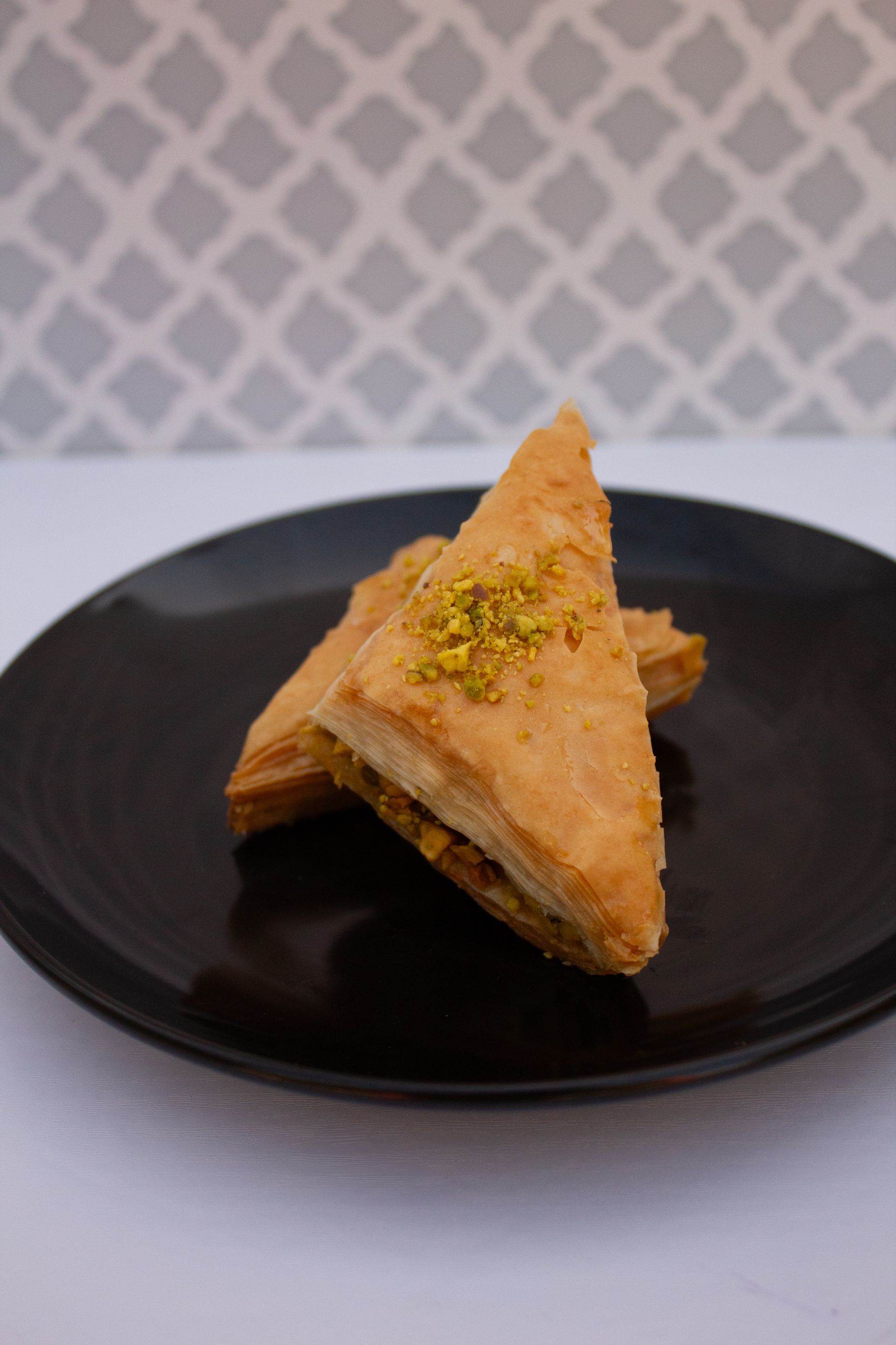 Two triangle-shaped pastries with pistachio topping on a black plate, against a patterned backdrop.