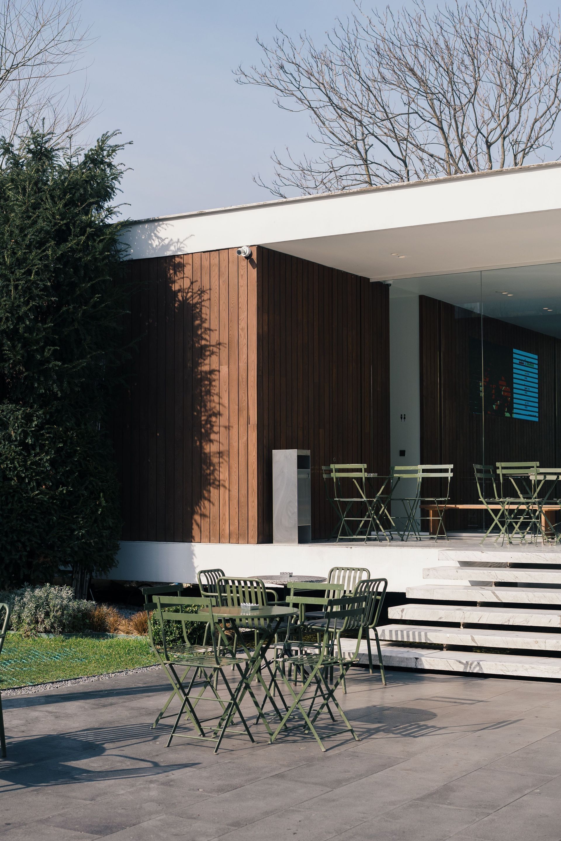 Outdoor café patio with green tables and chairs beside a modern building entrance