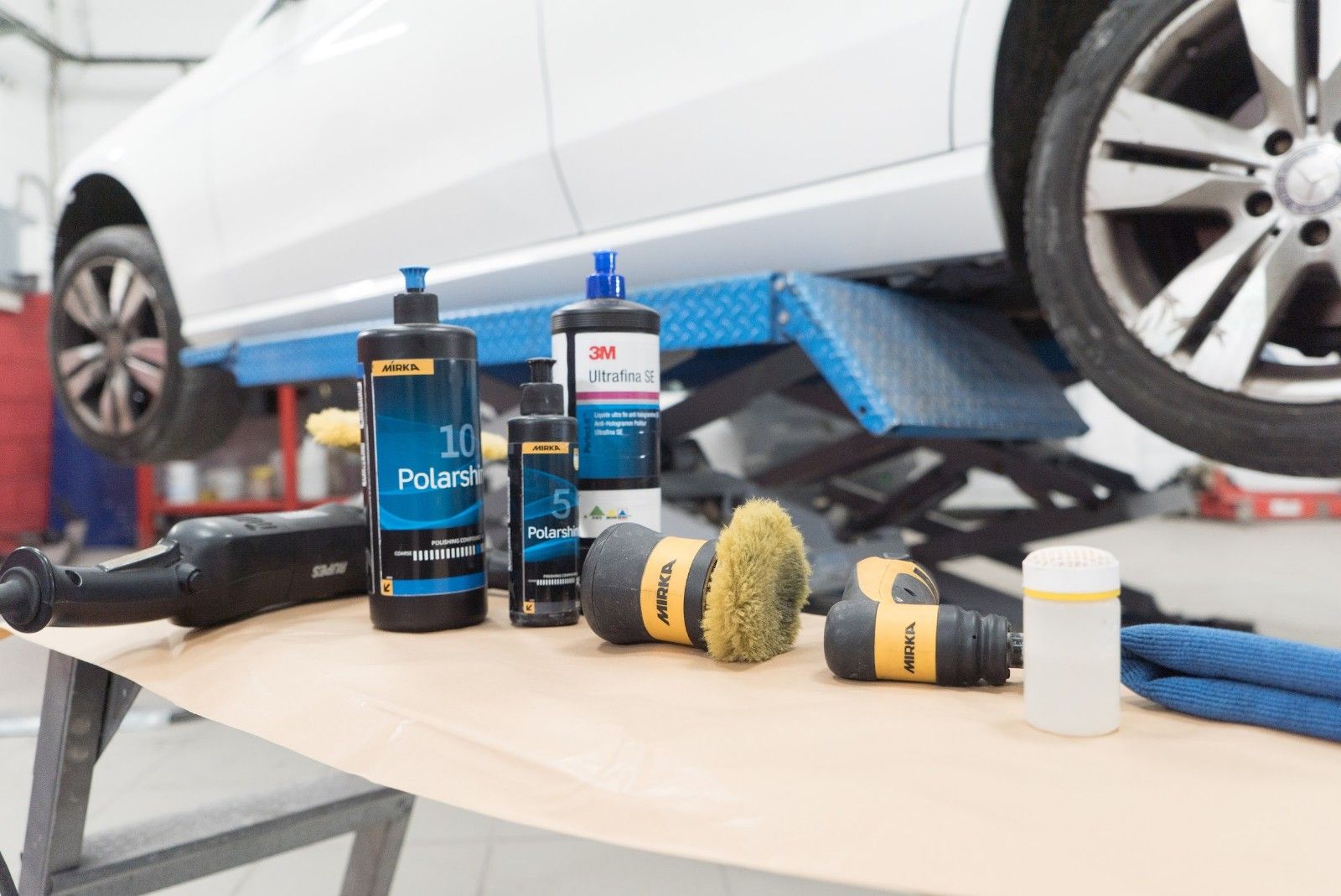 Un'auto bianca è parcheggiata su un ponte sollevatore in un garage, accanto a un tavolo con prodotti per la lucidatura.