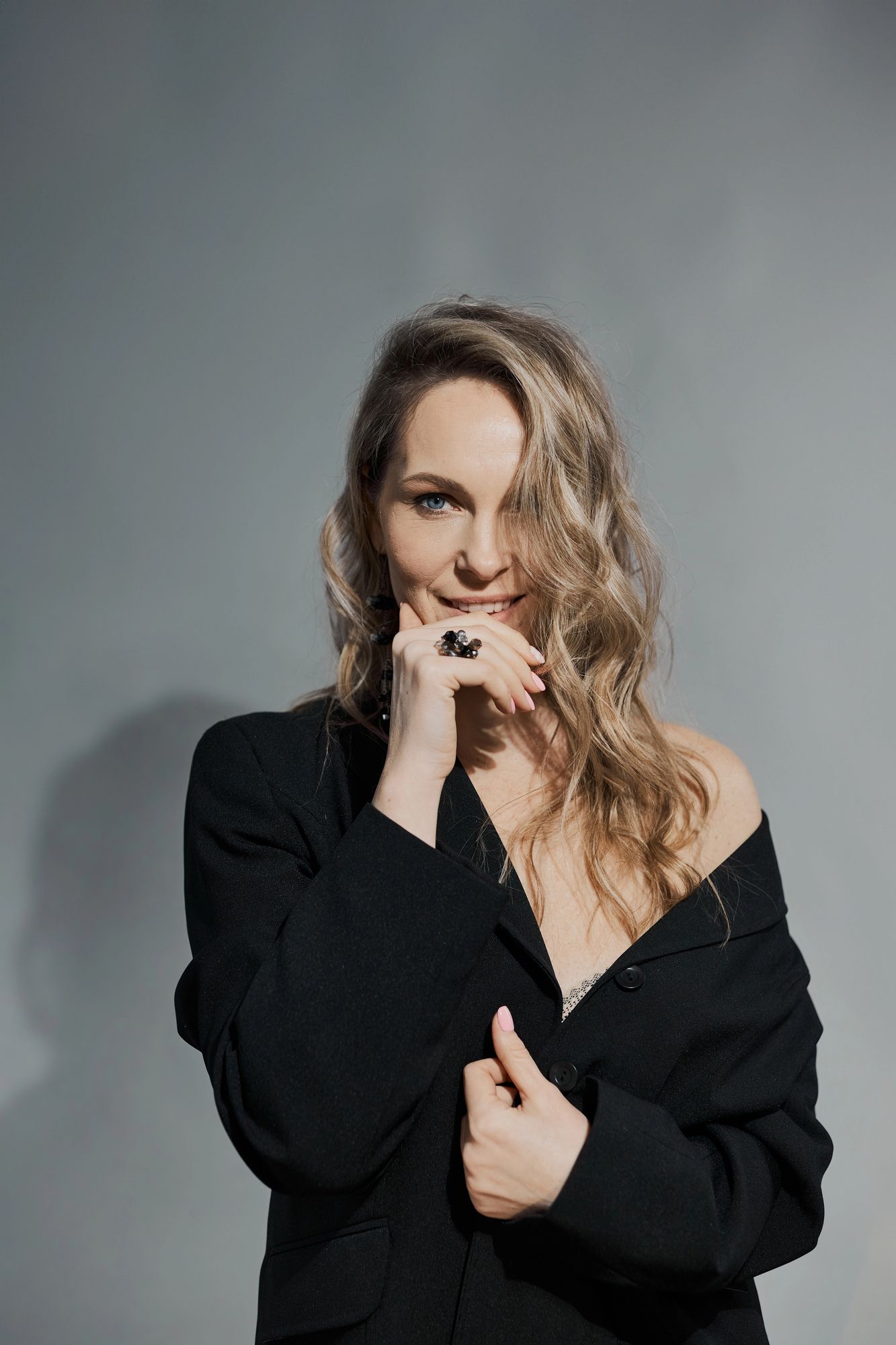 Donna con blazer nero, mano vicino alla bocca, sorridente, che guarda la telecamera su sfondo grigio.