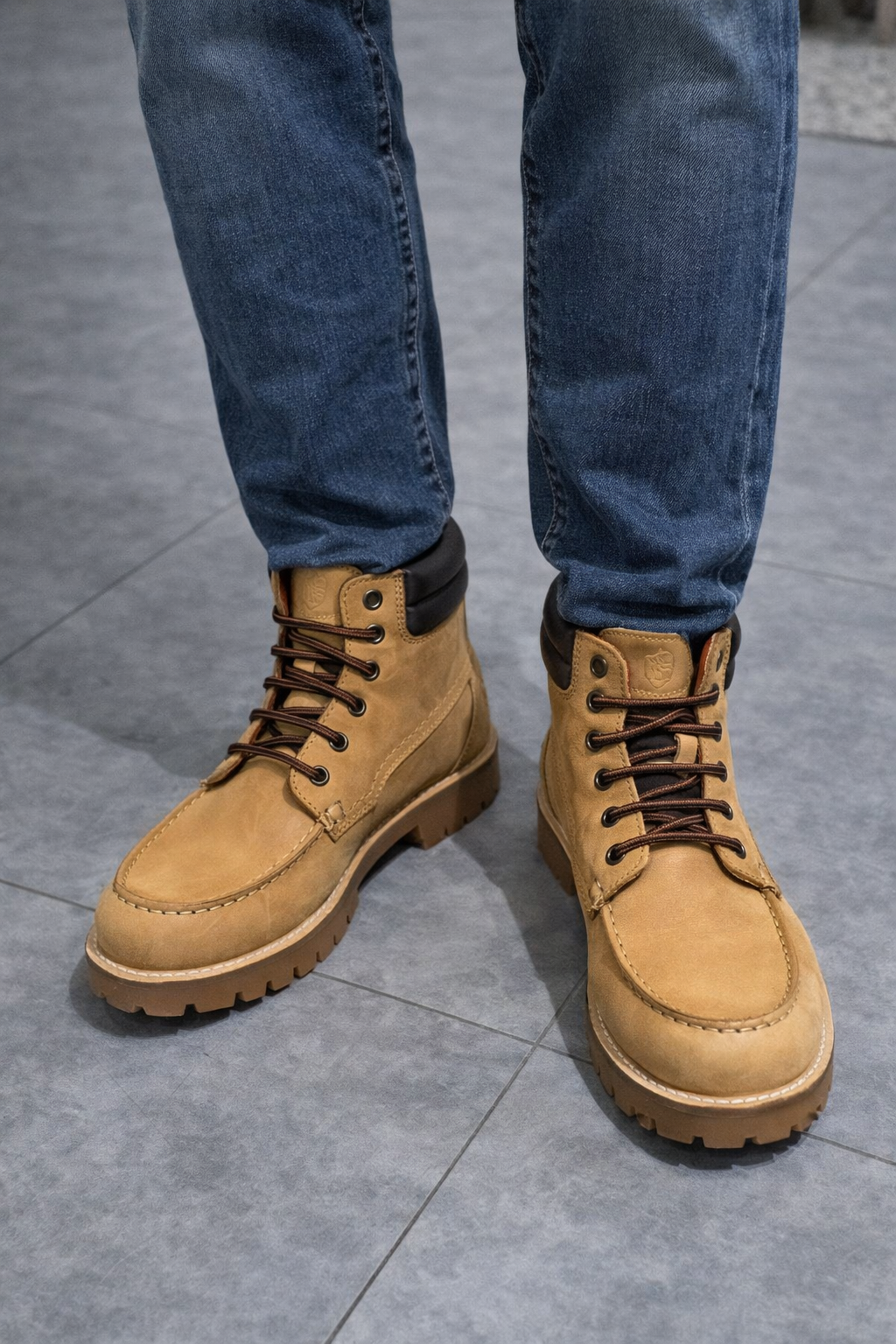 Stivali da lavoro color cuoio e jeans blu su un pavimento di piastrelle grigie.