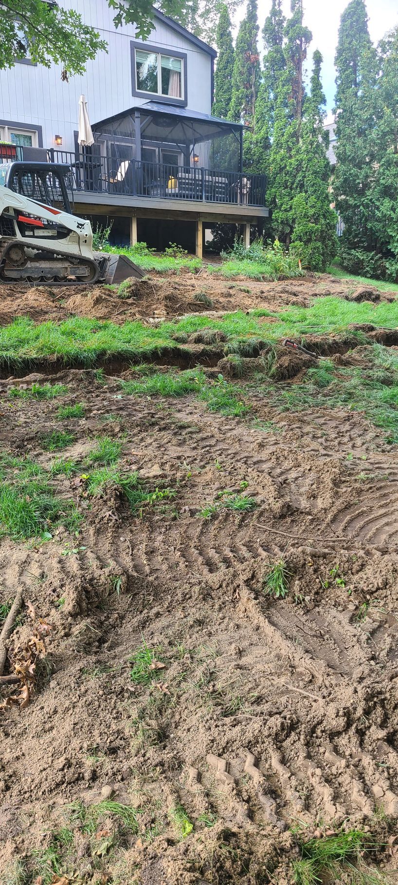 Backyard with excavated dirt, grass, and a deck with a screened porch in front of a house.