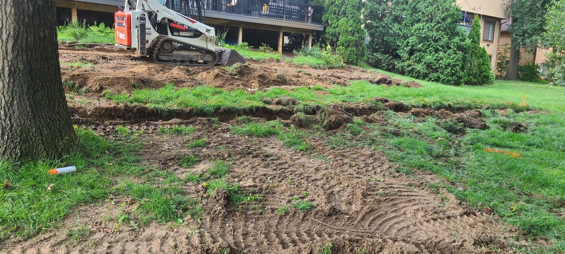 A small excavator digs into a sloped yard near a deck and trees.