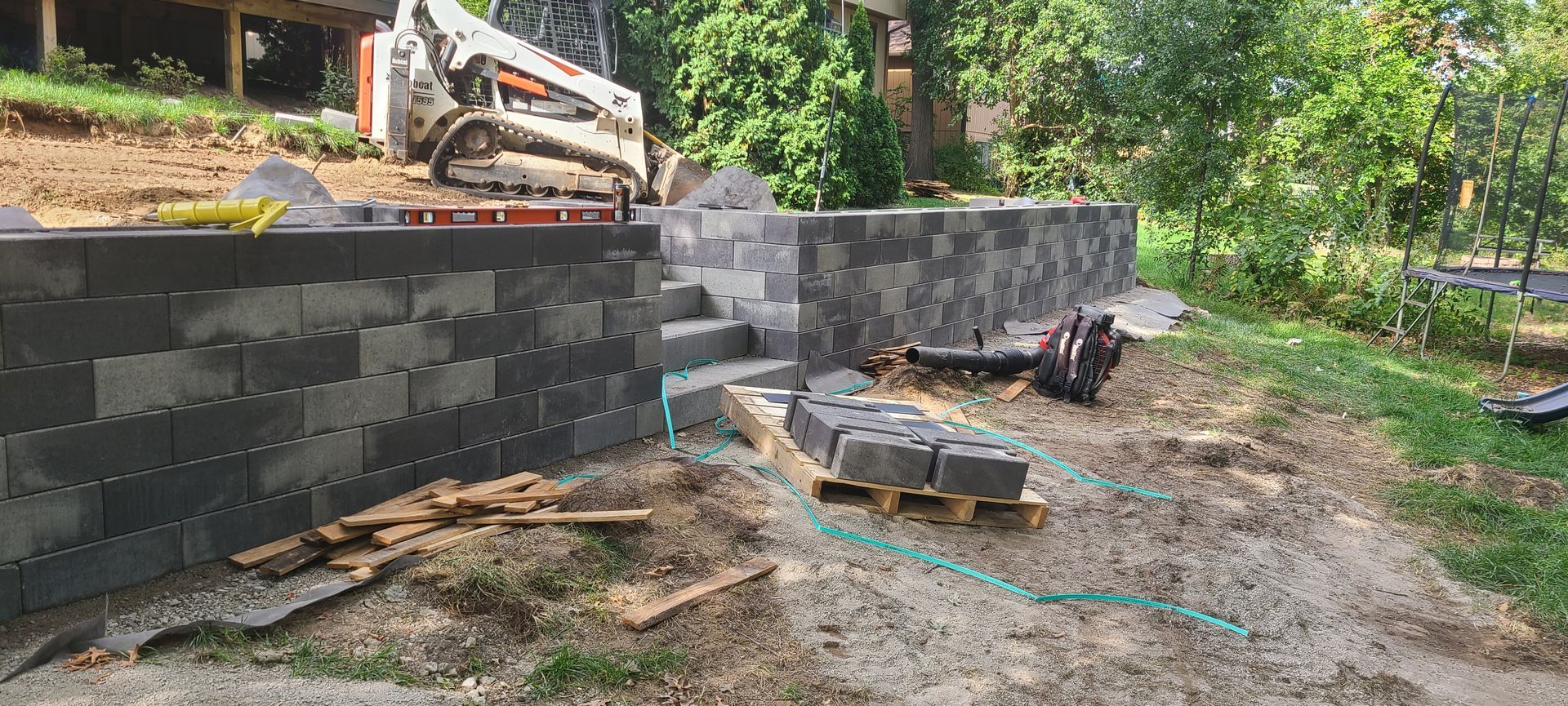 A retaining wall under construction with gray blocks. A skid steer loader is on the top, with greenery in the background.