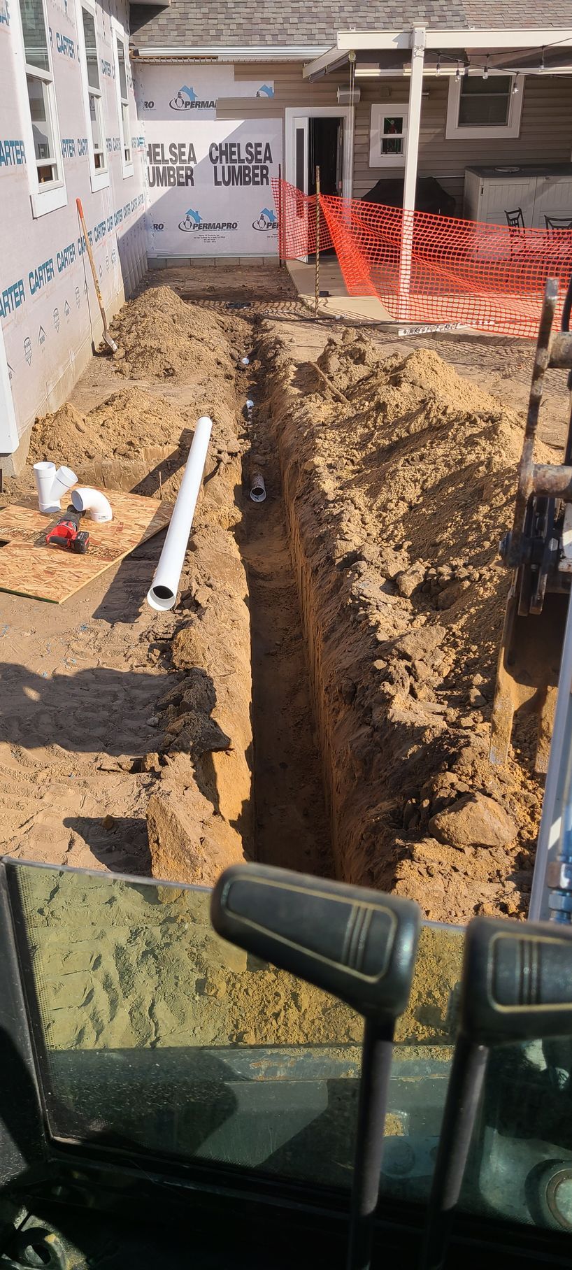 A long trench dug in dirt, with a white pipe inside and construction in the background.
