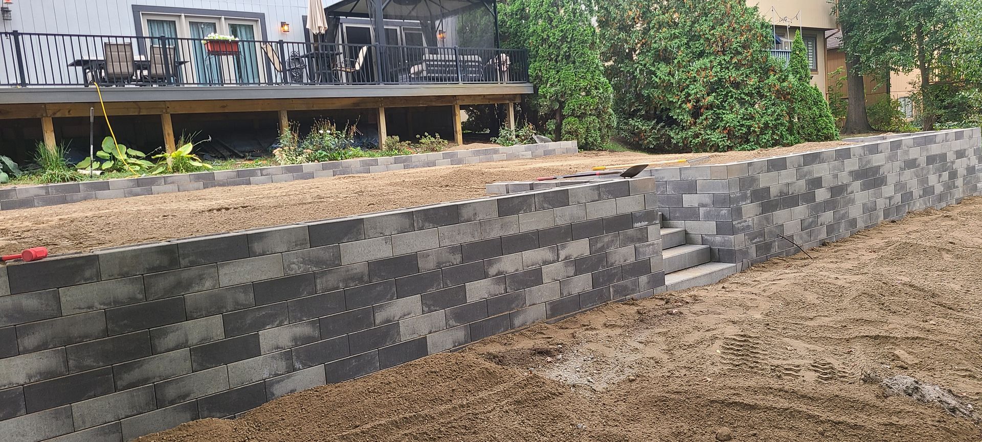 A gray retaining wall with steps leads to a deck. Dry dirt is in front of the wall.