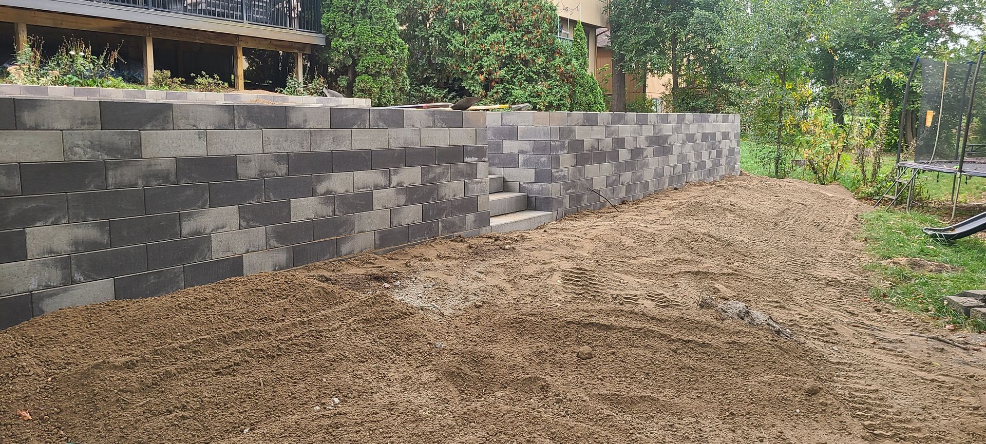 A retaining wall constructed of gray blocks with brown soil in front. Green trees in the background.