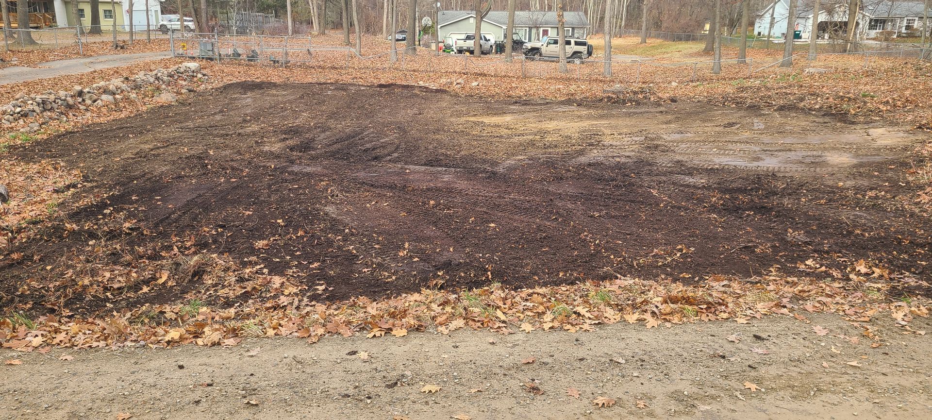 A freshly tilled garden plot covered with dark soil and fallen leaves. Trees and distant buildings in the background.