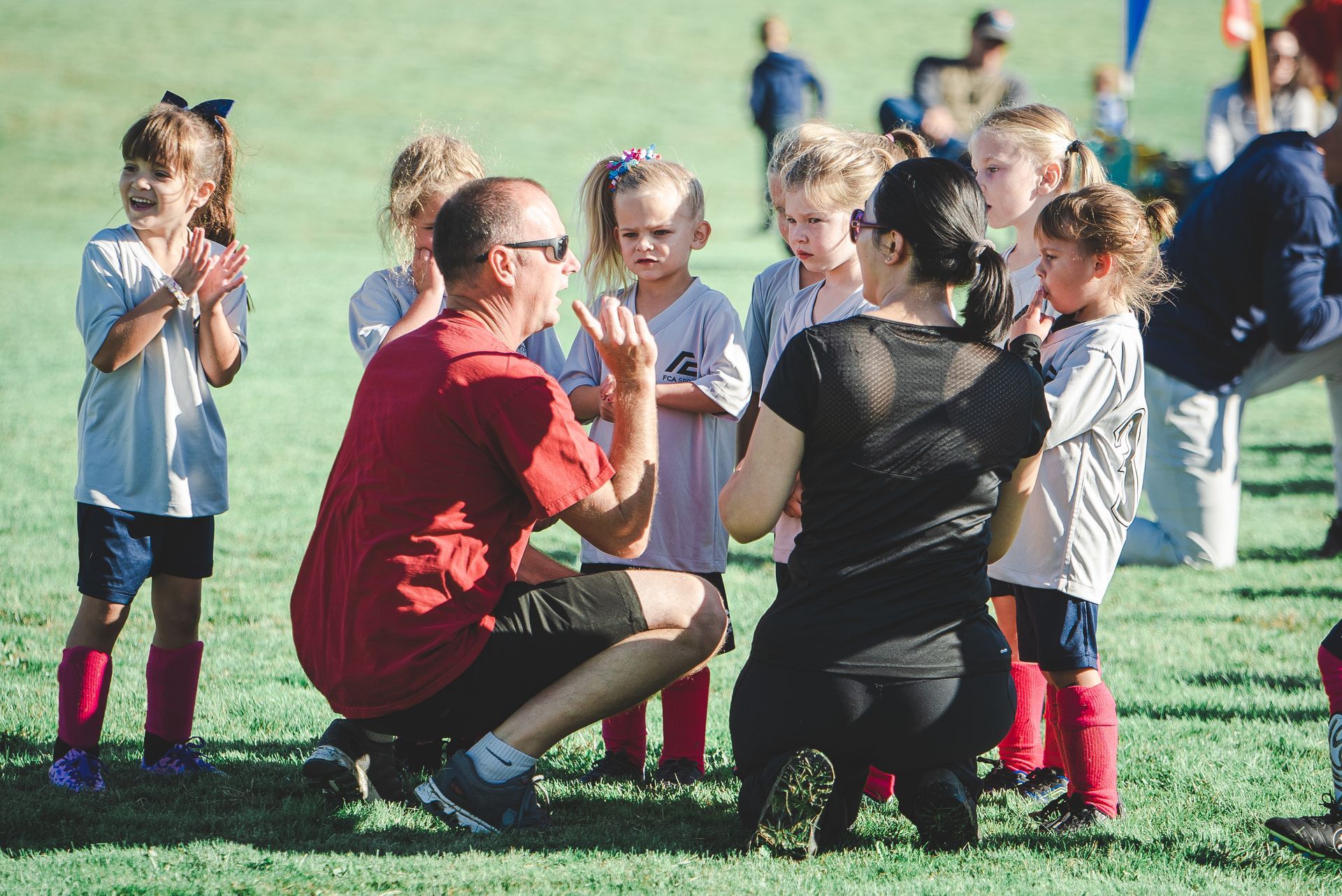 Coach kneels with children on a grassy field during an outdoor sports practice.