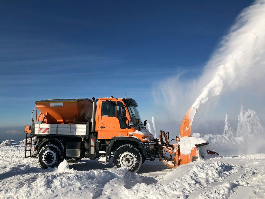 Orange snowplow clearing deep snow with a plume of white snow under a clear blue sky