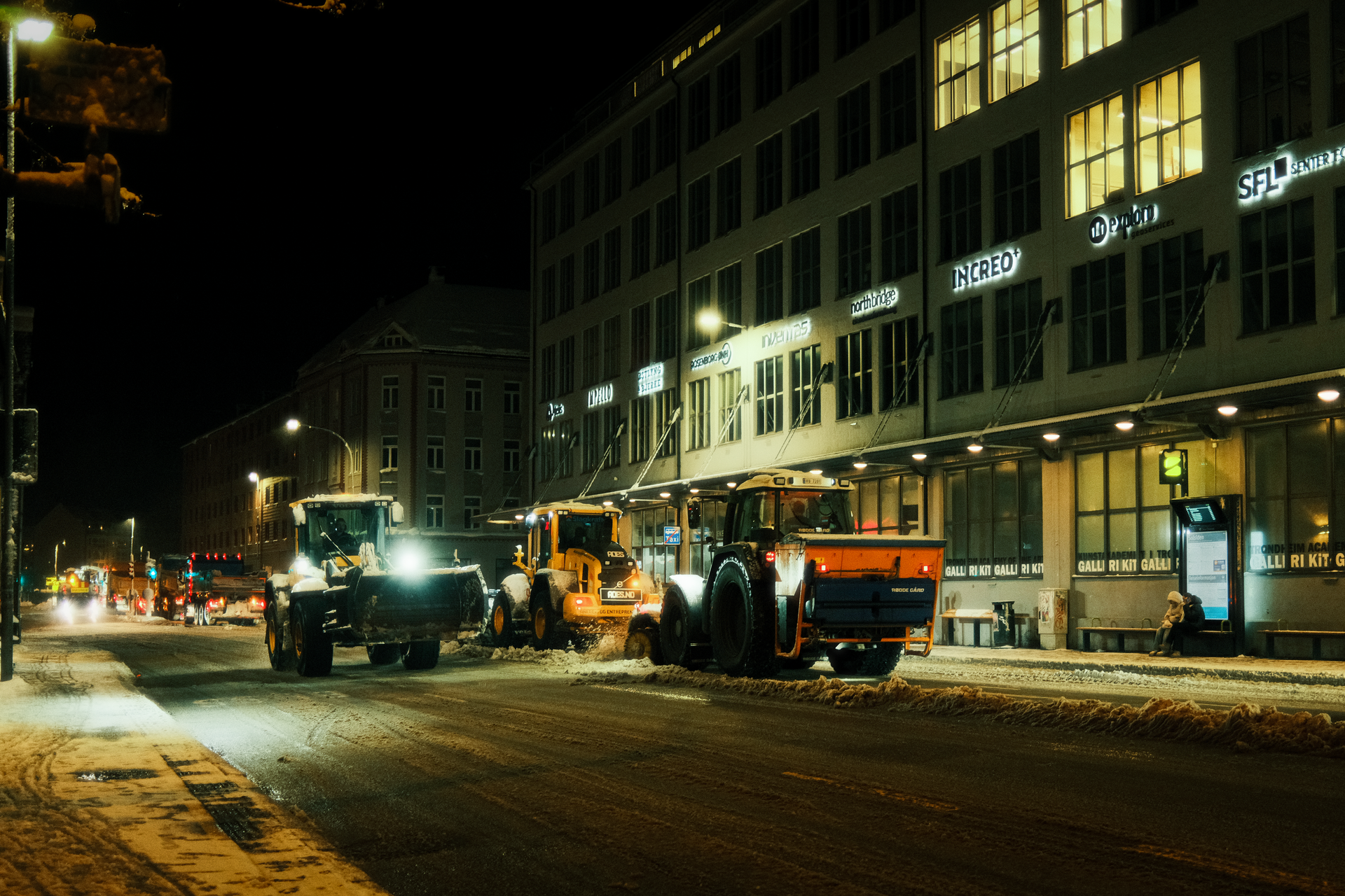 Snowplows clearing a snowy city street at night beside lit storefronts and buildings