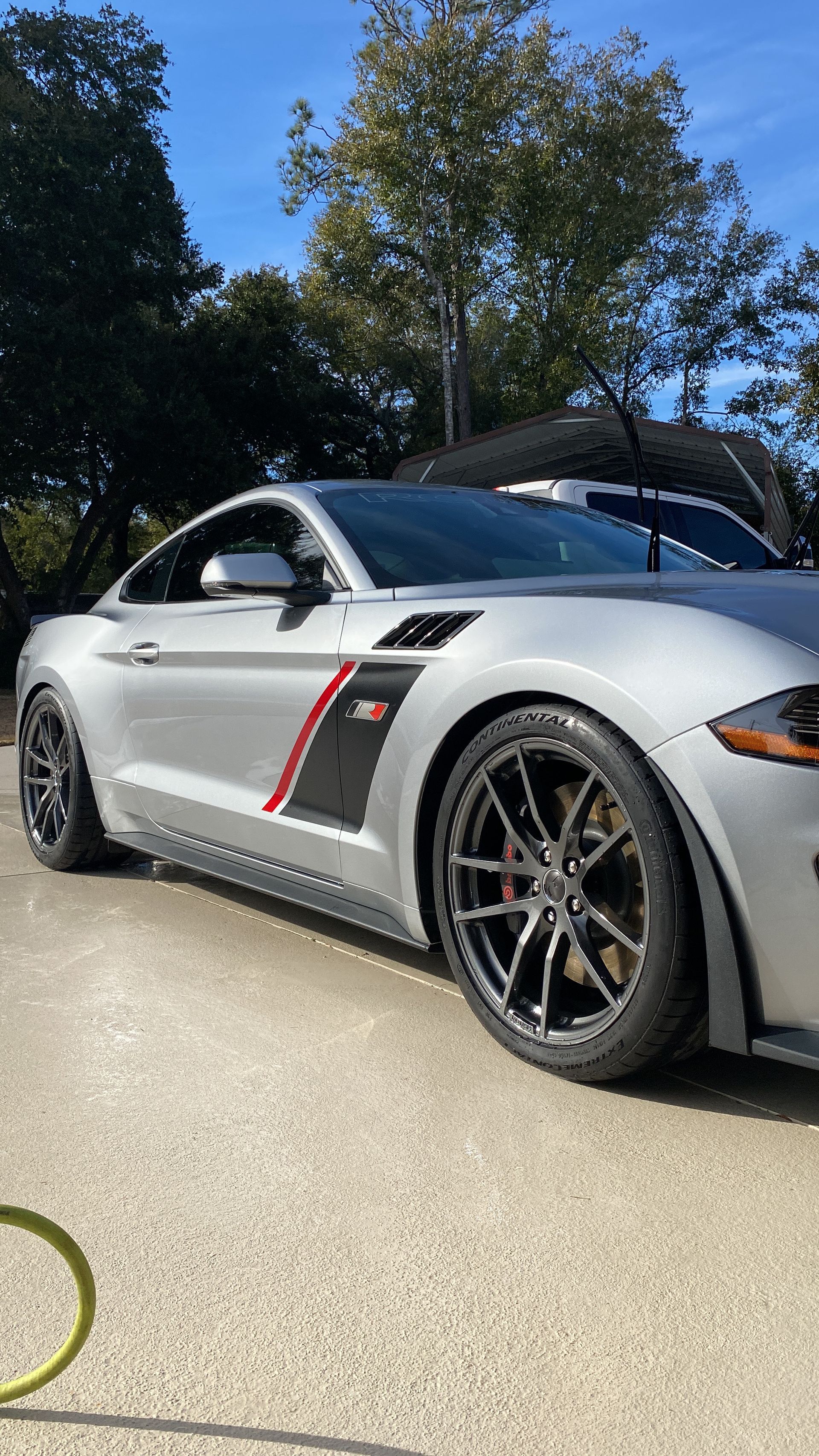 A silver mustang is parked in a driveway with trees in the background.