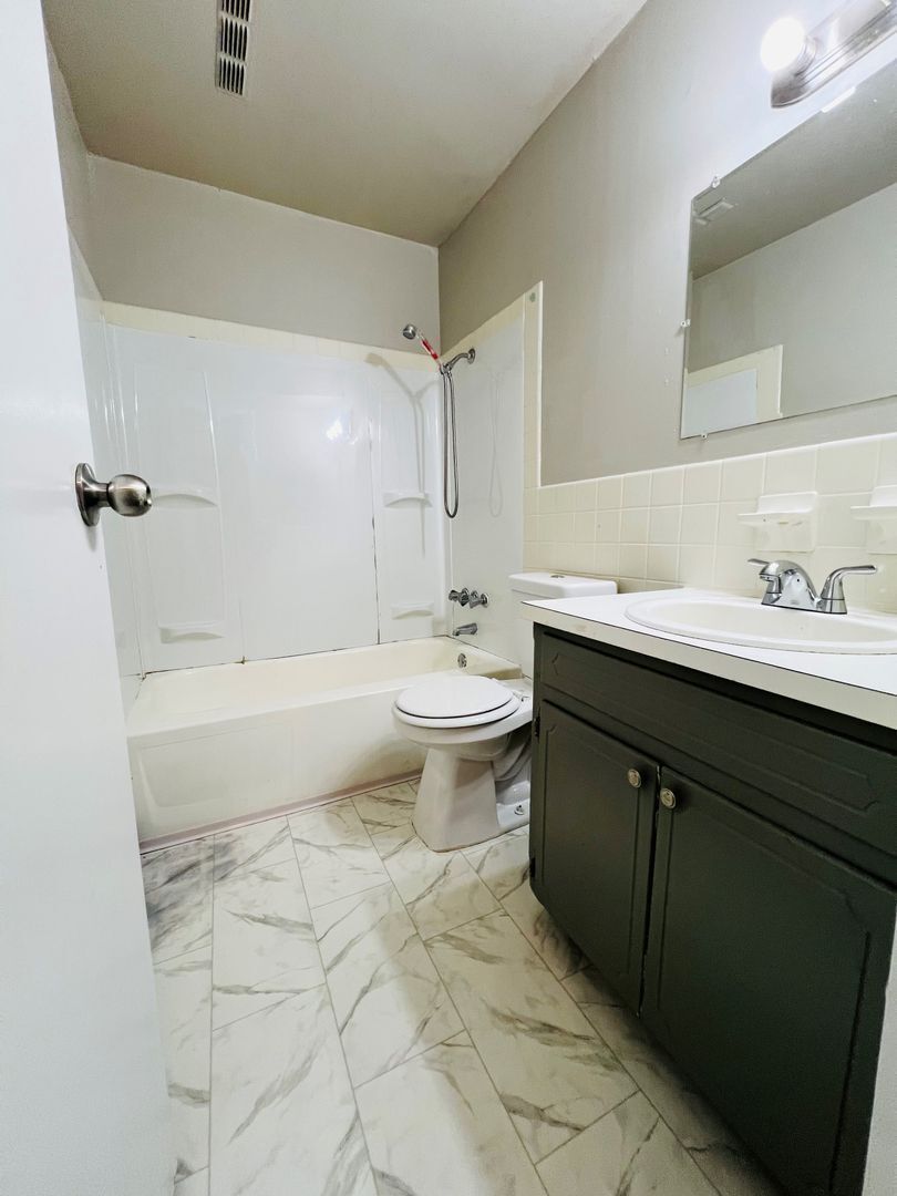 Small bathroom with white tub, toilet, vanity, mirror, and marble-pattern floor.