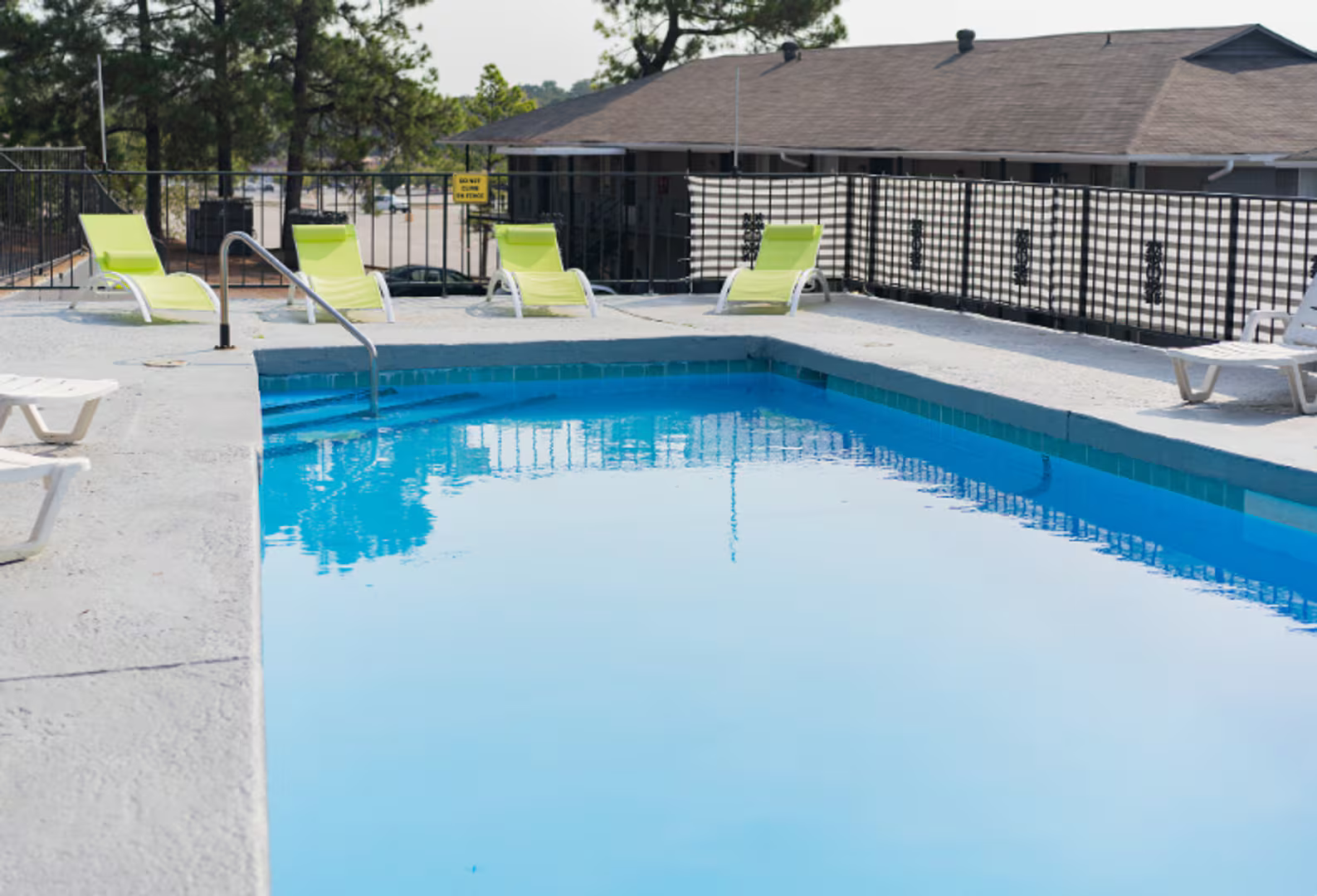 Outdoor swimming pool with blue water, white deck chairs, and a fenced building in the background